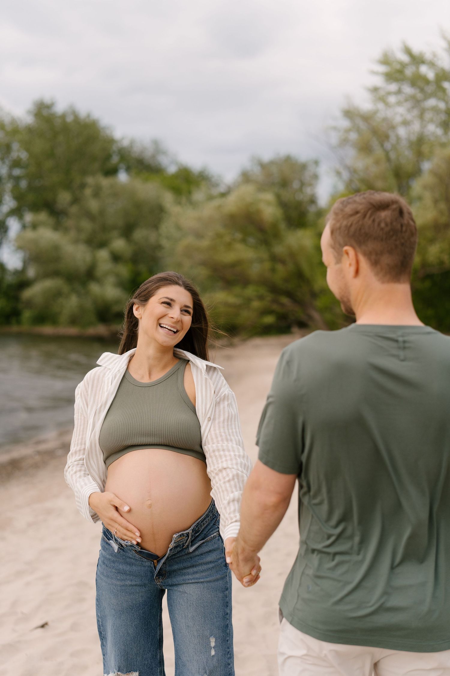 Laughing pregnant woman in green top and jeans holds belly while holding hands with partner on beach.