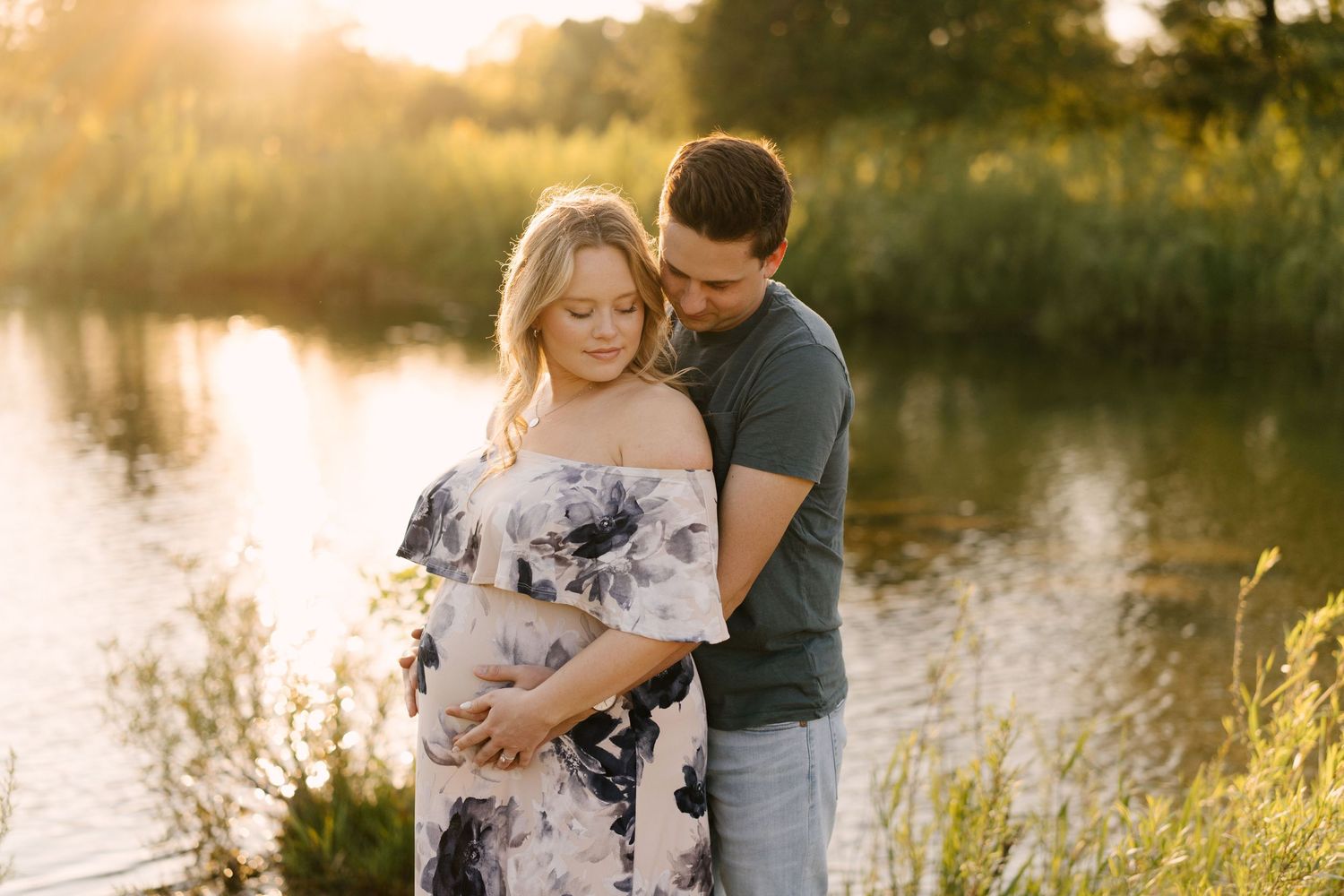 Couple embraces tenderly by a glowing sunset lake, hands resting on pregnant woman's floral dress belly.