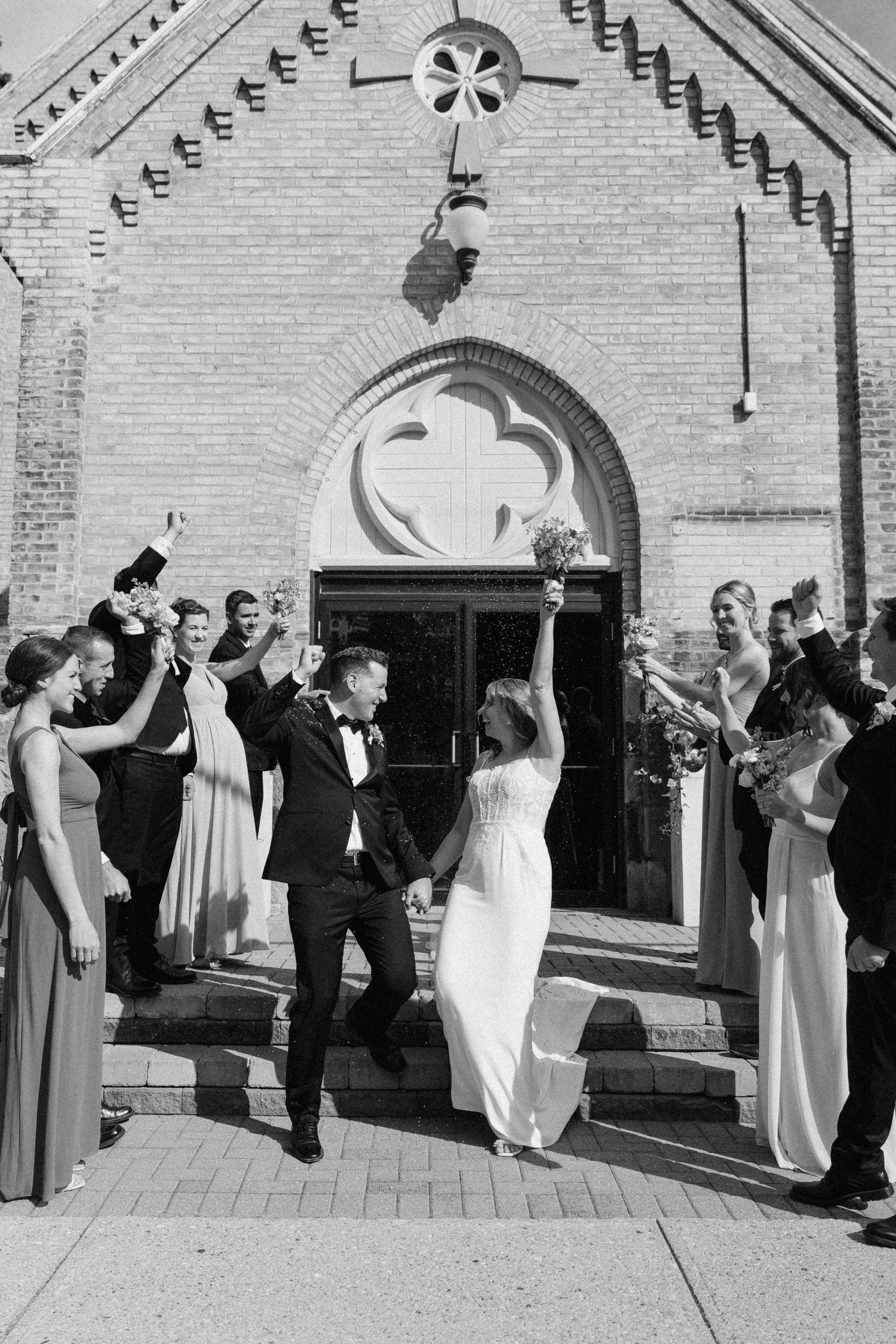 Black and white photo of joyful newlyweds exiting a brick church, surrounded by cheering wedding party.