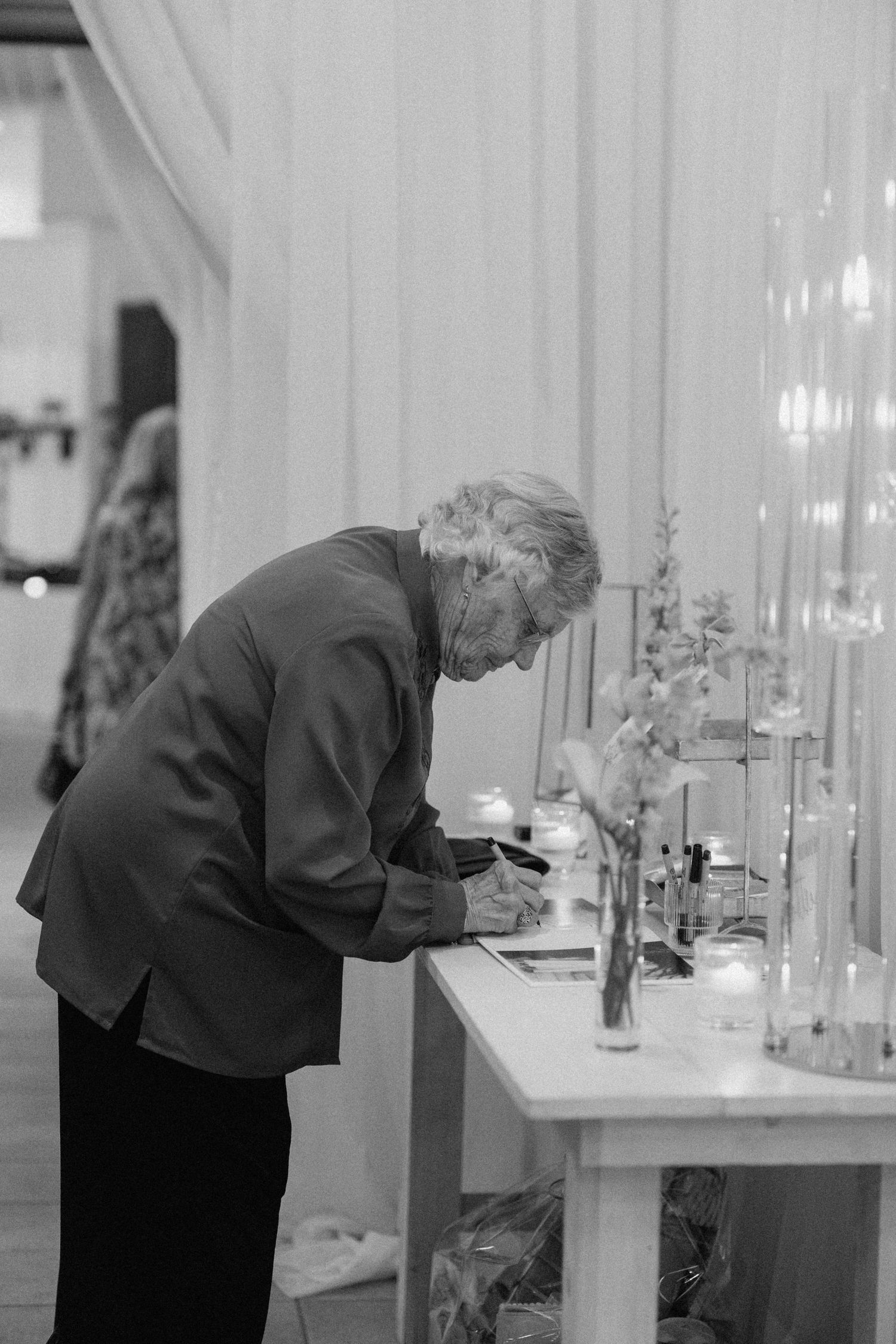 Black and white photo of elderly guest signing wedding guestbook at floral decorated table.