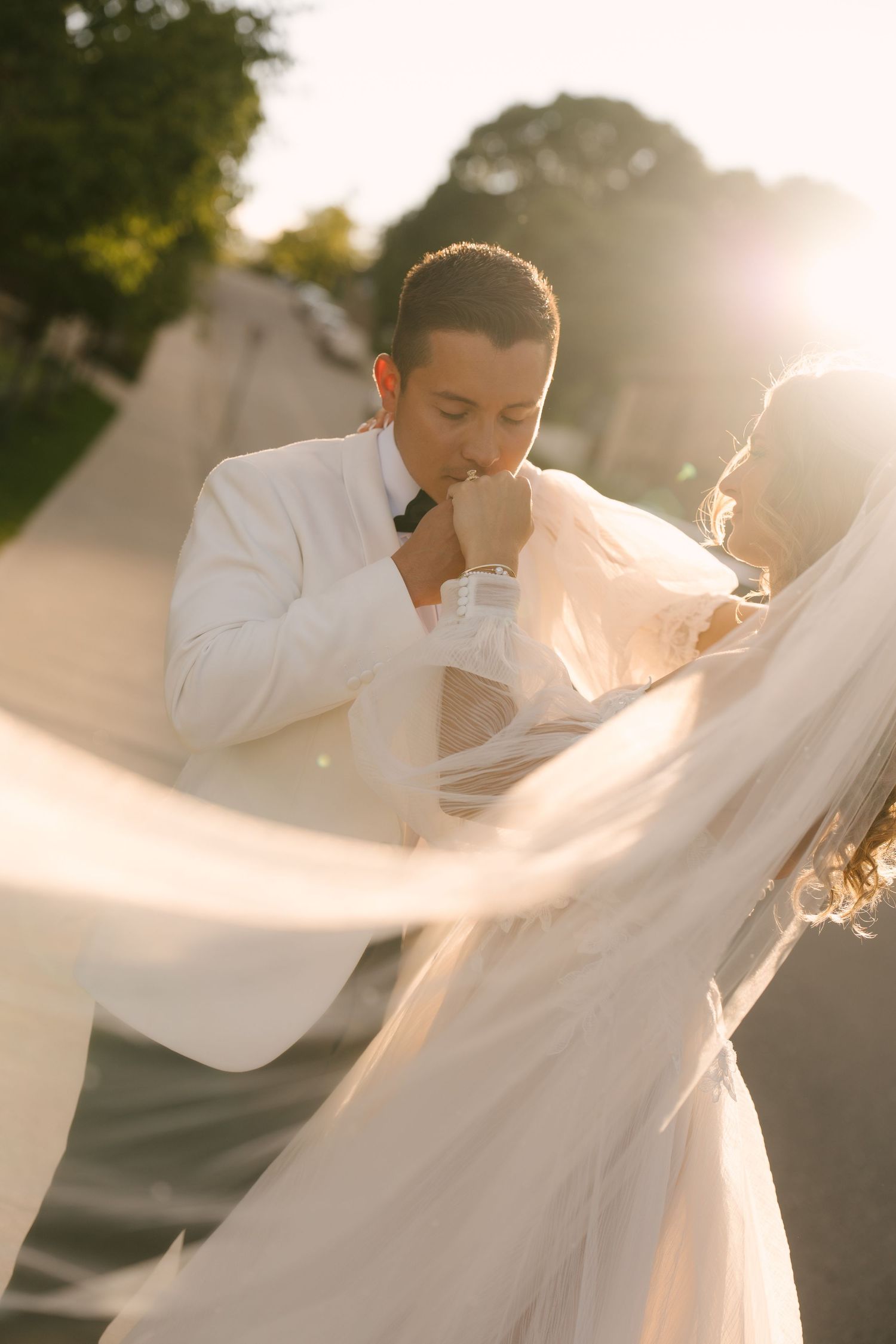 Groom in white suit kissing bride's hand with veil flowing in golden sunlight outside Tapestry Hall wedding venue.