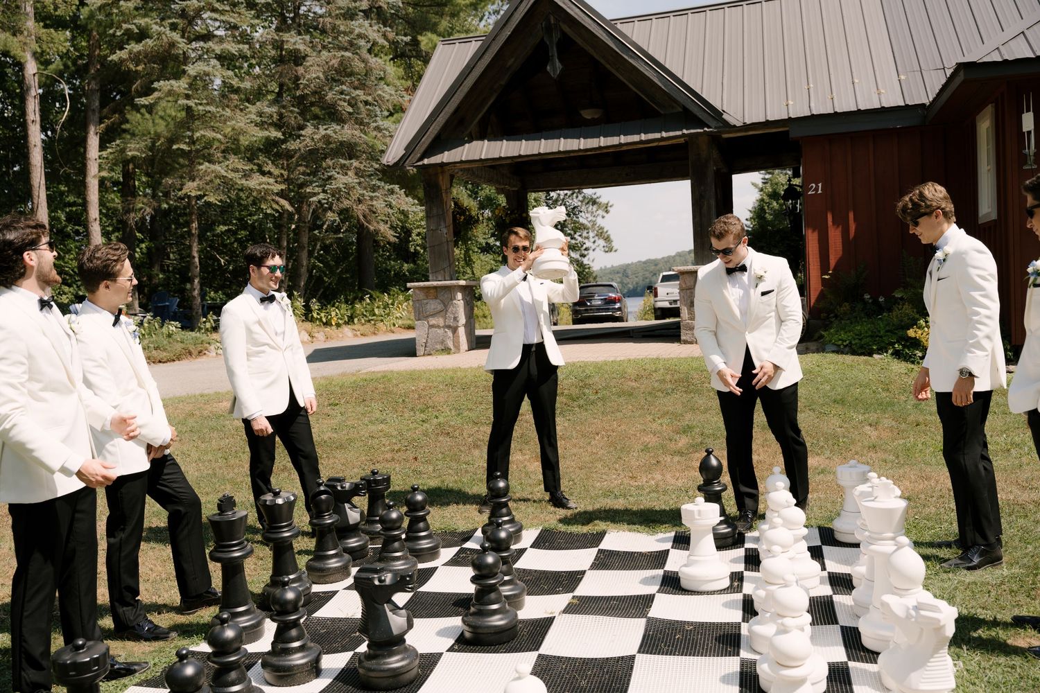 Groomsmen in white tuxedo jackets play giant outdoor chess on a black and white board at a wedding venue.