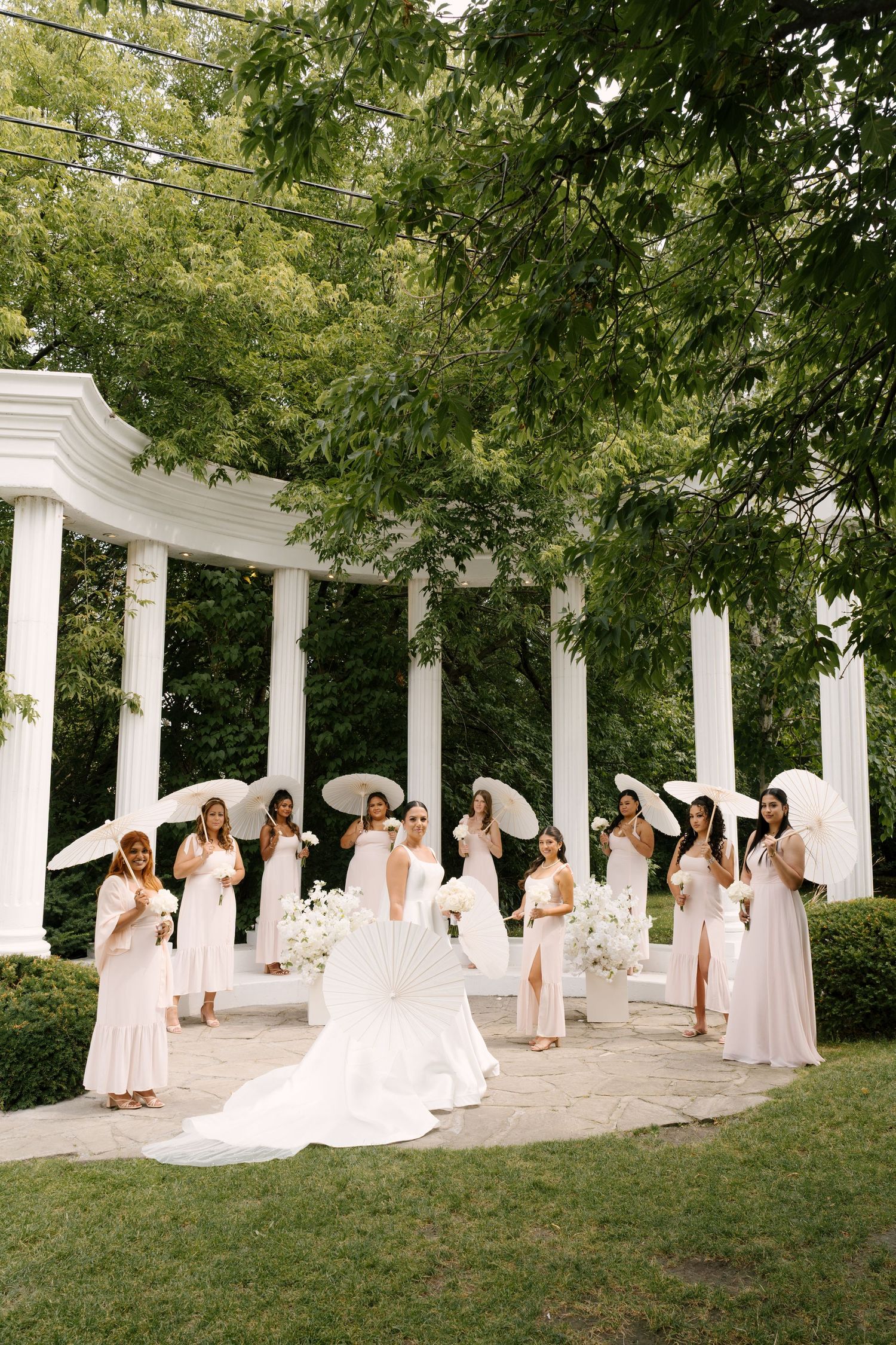 Bride surrounded by bridesmaids in blush dresses holding white parasols near a white columned garden pavilion.