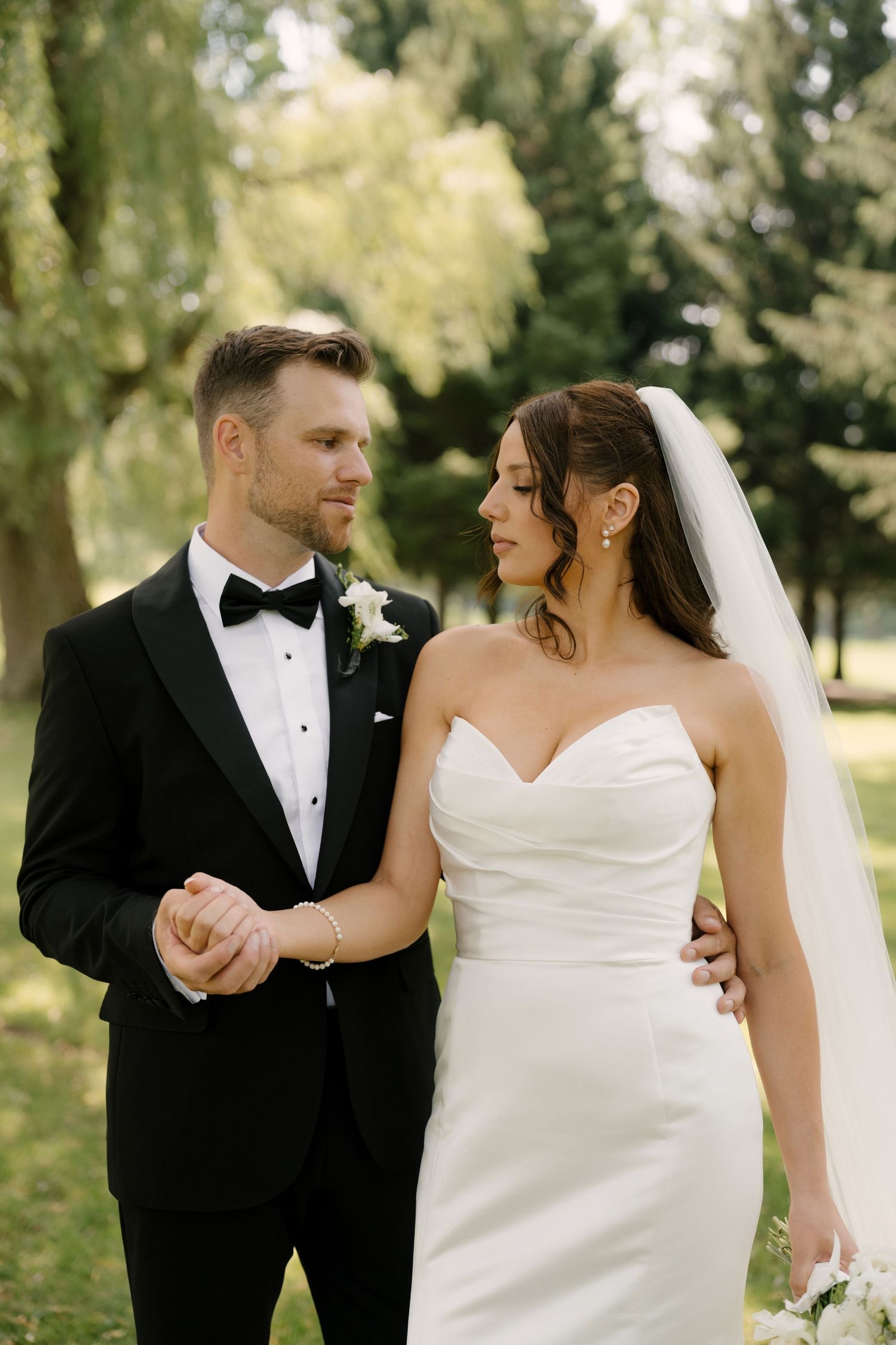 Bride and groom share a romantic moment outdoors, she in a strapless white gown with veil, he in a black tuxedo.