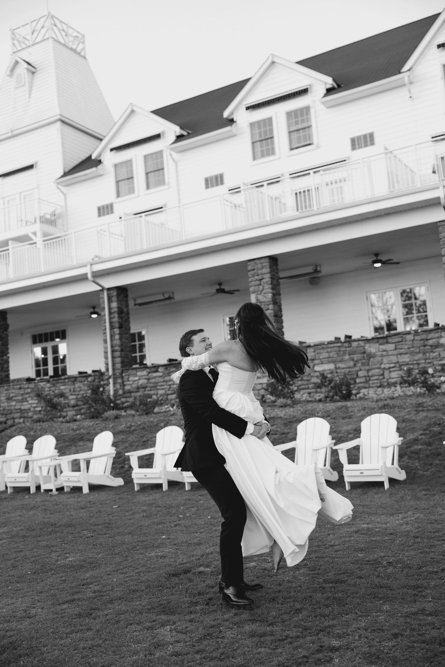 Black and white photo of groom lifting and spinning bride on lawn in front of elegant venue.