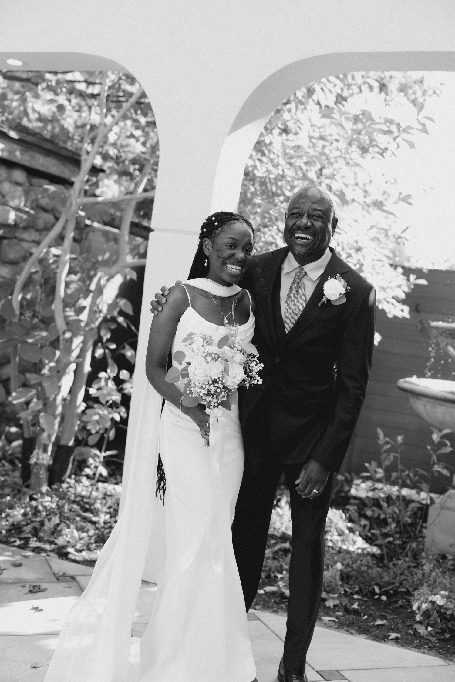 A smiling bride holding a bouquet poses with a laughing older man under a white arch in a black and white photo at Wren House venue.