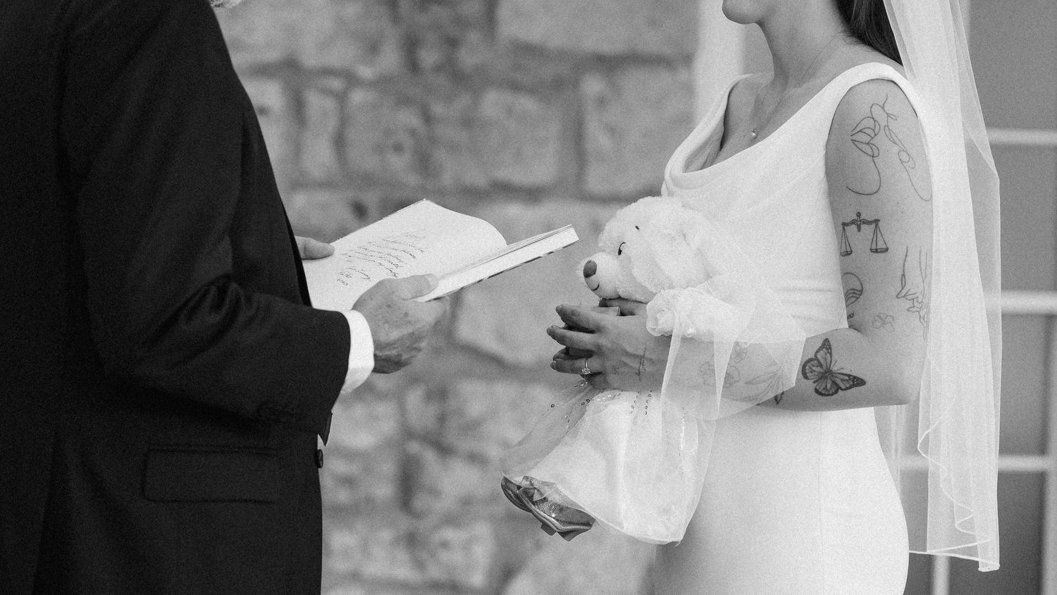 Bride holding a stuffed teddy bear while exchanging vows during outdoor wedding ceremony.