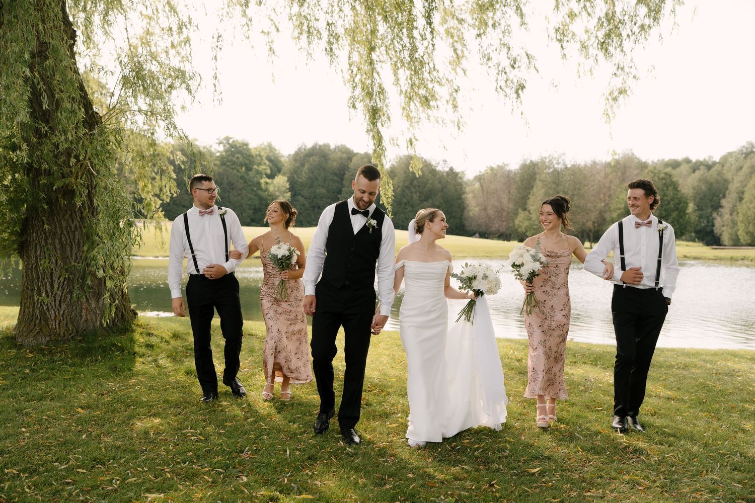 Wedding party walks together near scenic pond under weeping willow tree on sunny day at Guelph golf club.
