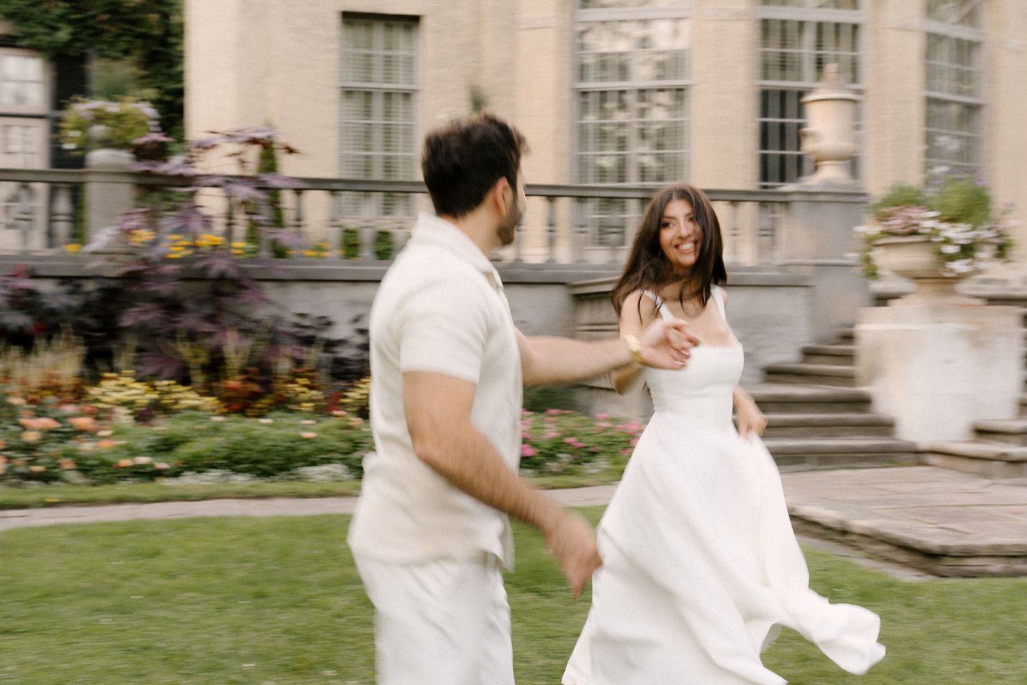 Couple in white outfits dancing joyfully on a lush garden lawn near a grand estate building.