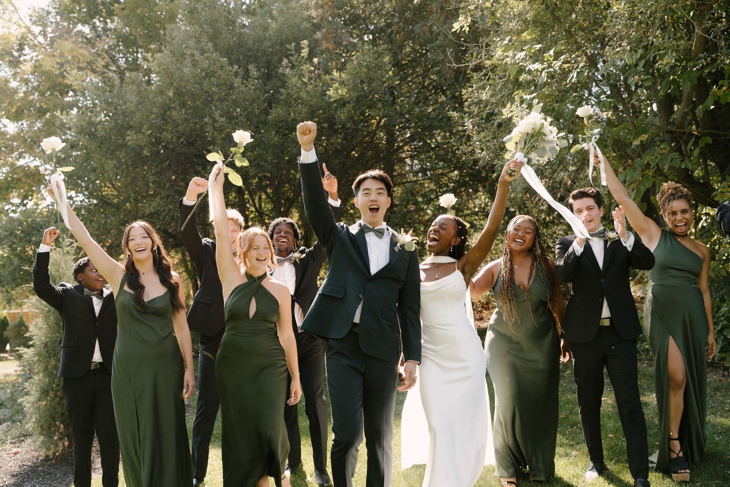 Joyful wedding party celebrating with raised arms and bouquets in sunlit outdoor setting at Wren House.