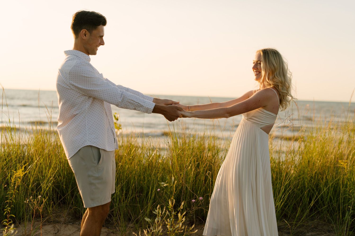 Couple holding hands in tall grass near beach at sunset.