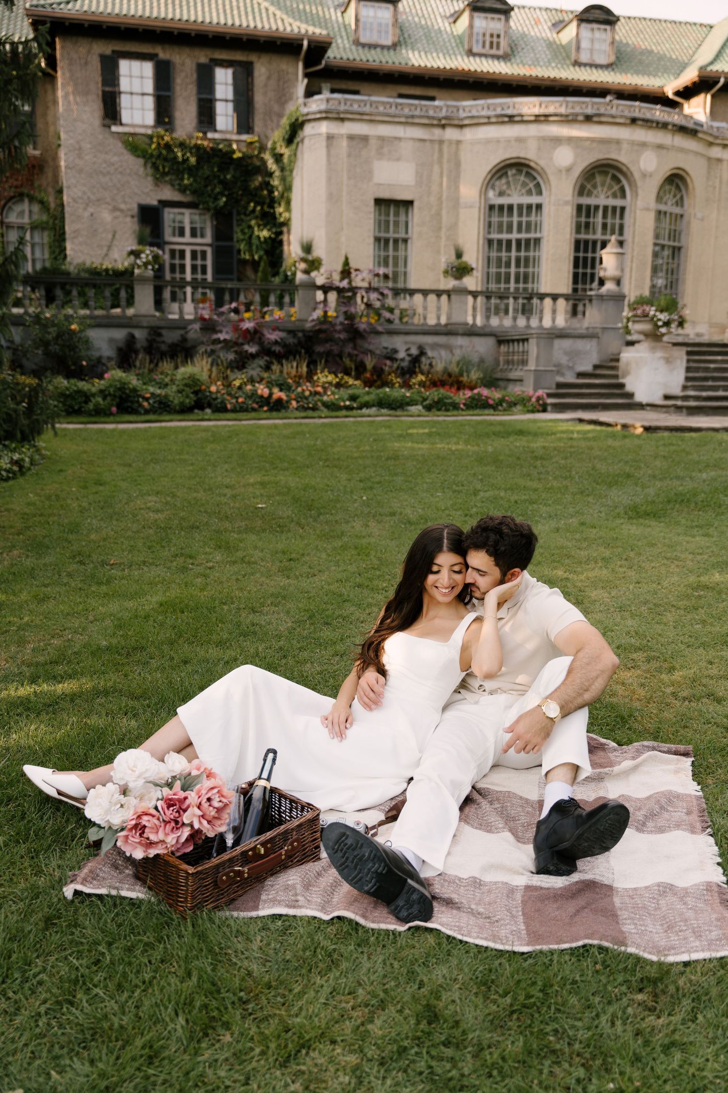 Couple sharing a tender moment on a picnic blanket with flowers and basket on a green lawn before a mansion.