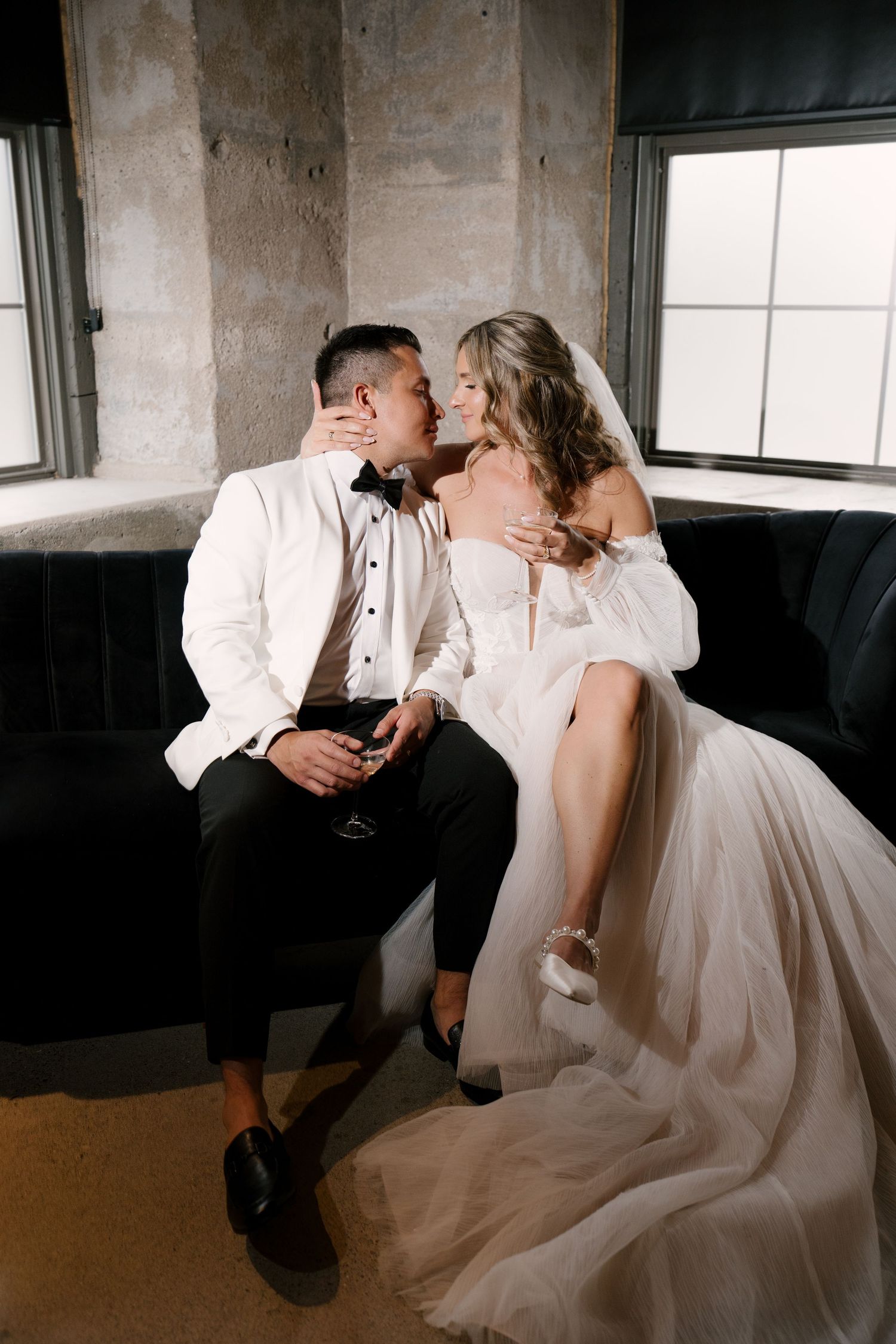 Bride in off-shoulder tulle gown sits on grooms lap as they share an intimate moment on a dark velvet sofa at Tapestry Hall wedding venue.