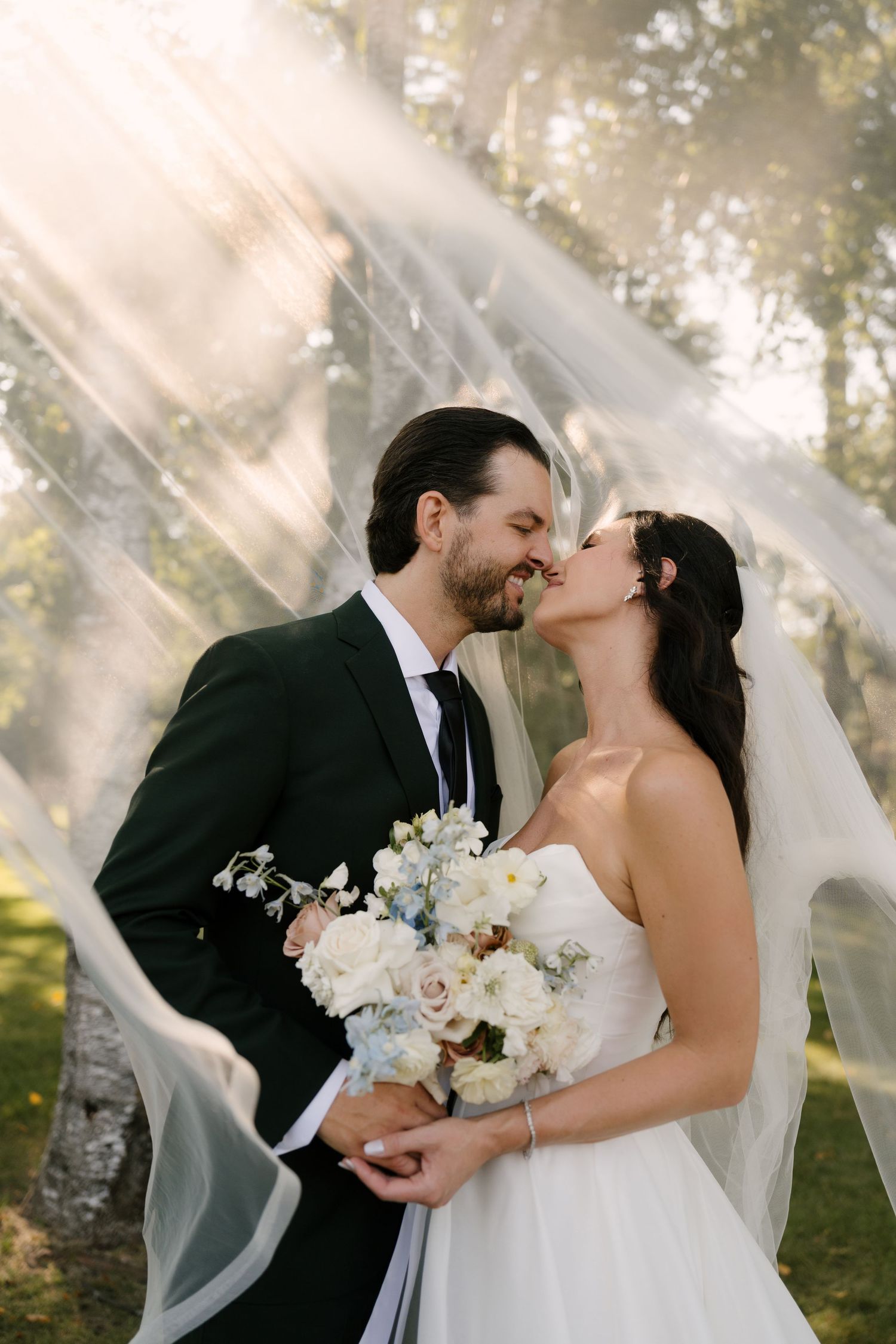 Bride and groom sharing a romantic kiss outdoors, bride holding white floral bouquet, veil flowing in sunlight at Roseville Estates.