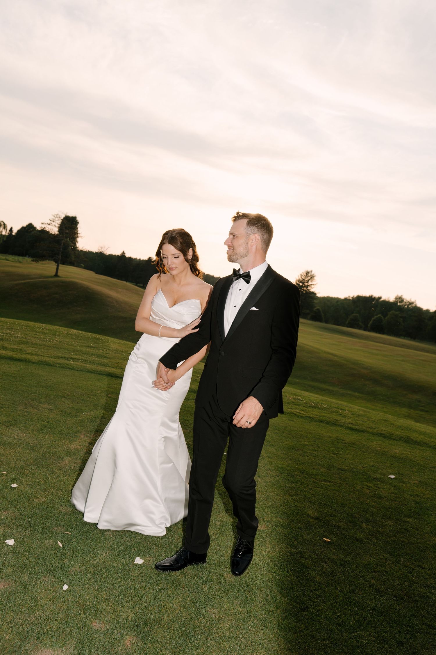 Bride and groom walking hand in hand across a green golf course at dusk in formal wedding attire.
