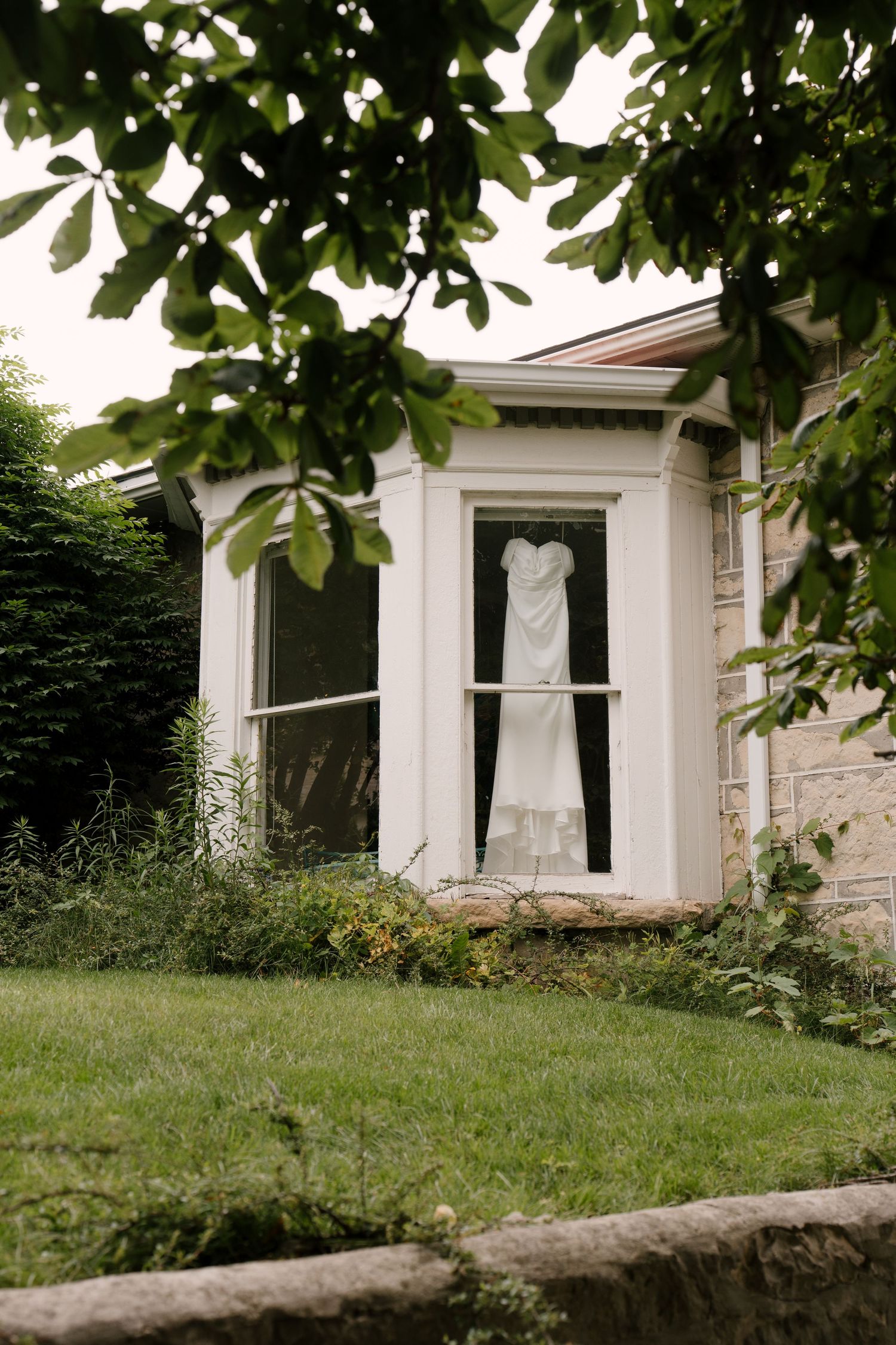 White wedding dress hanging in bay window of stone cottage surrounded by lush garden greenery.