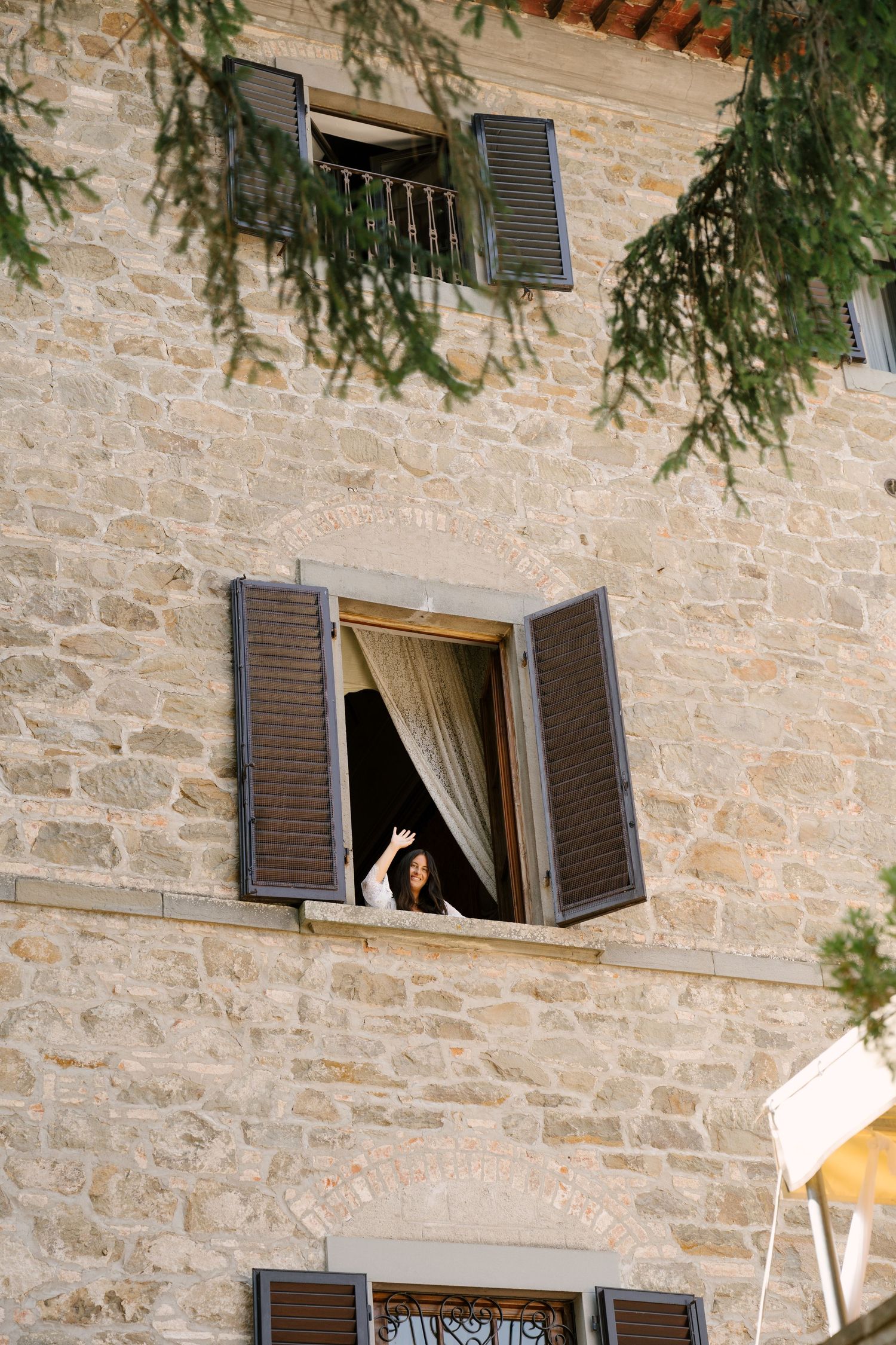 Bride waving from open window of rustic stone building with wooden shutters and cypress tree branches overhead.