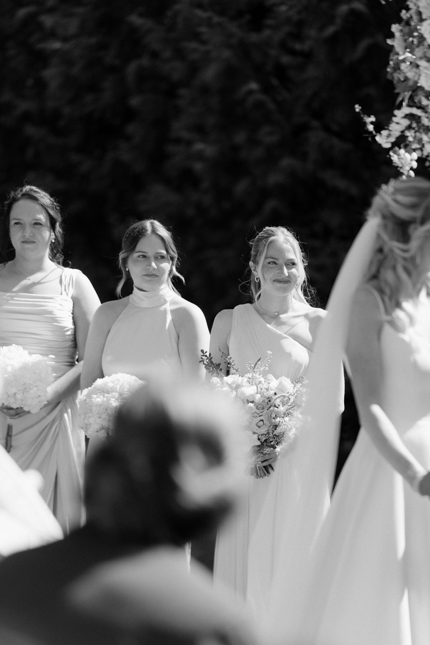 Bridesmaids in white gowns holding bouquets watch attentively during outdoor ceremony at Roseville Estates wedding venue.