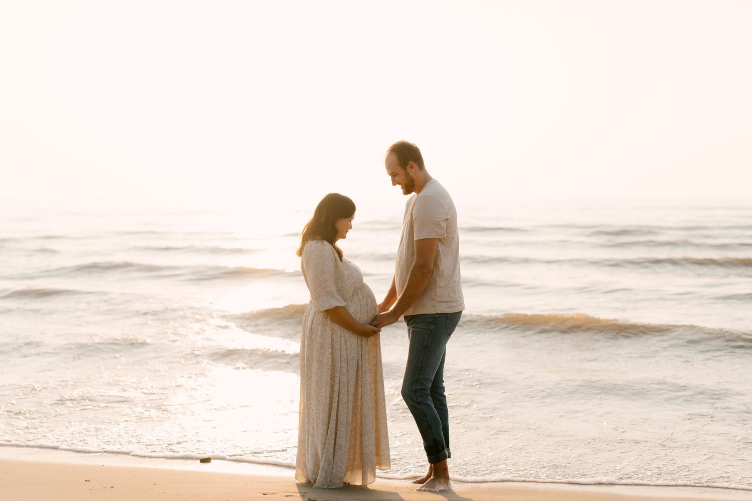 Pregnant couple standing together on beach at sunset, holding hands and gazing at each other.