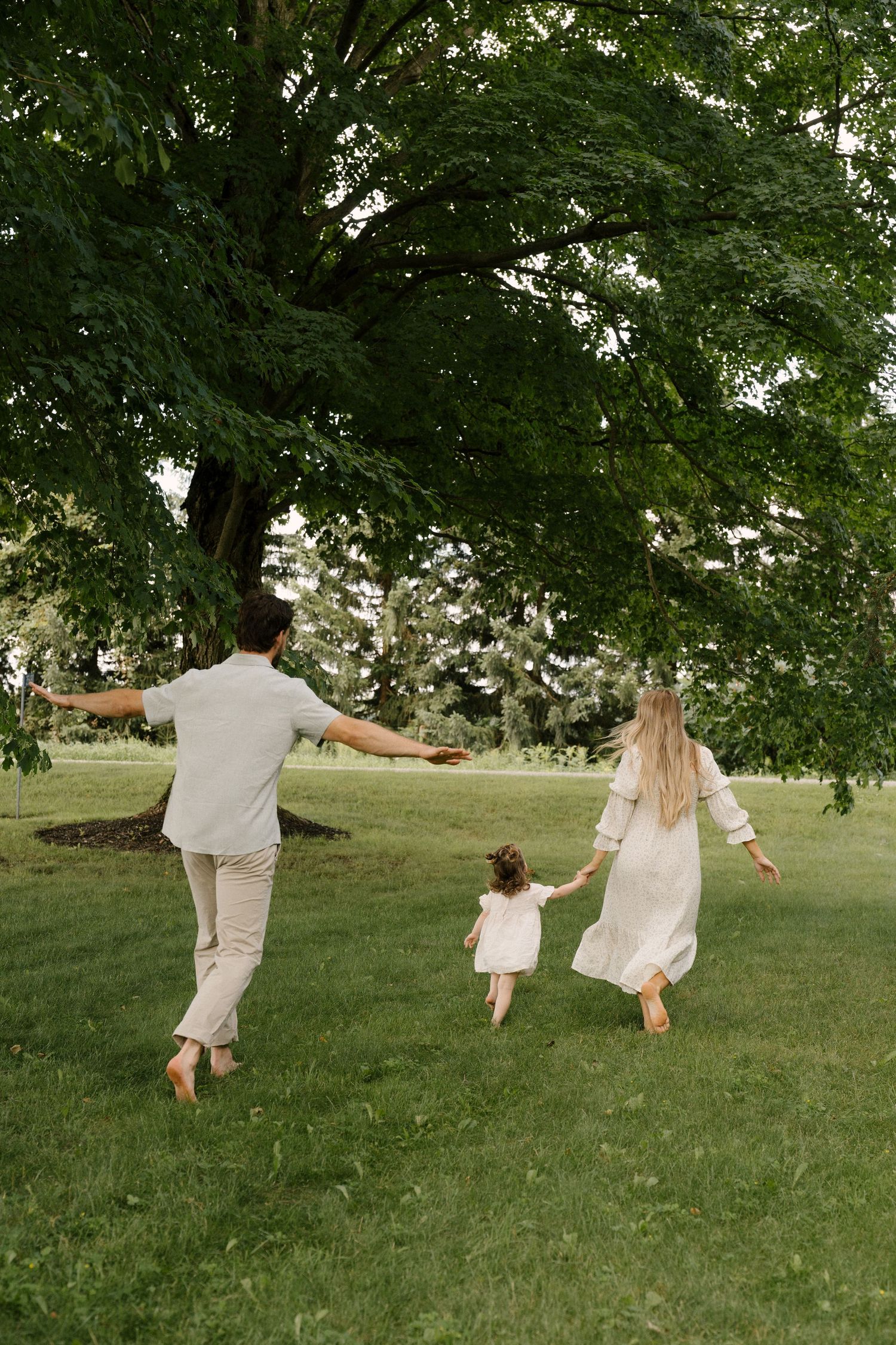Family of three holding hands walking barefoot toward large tree in park.