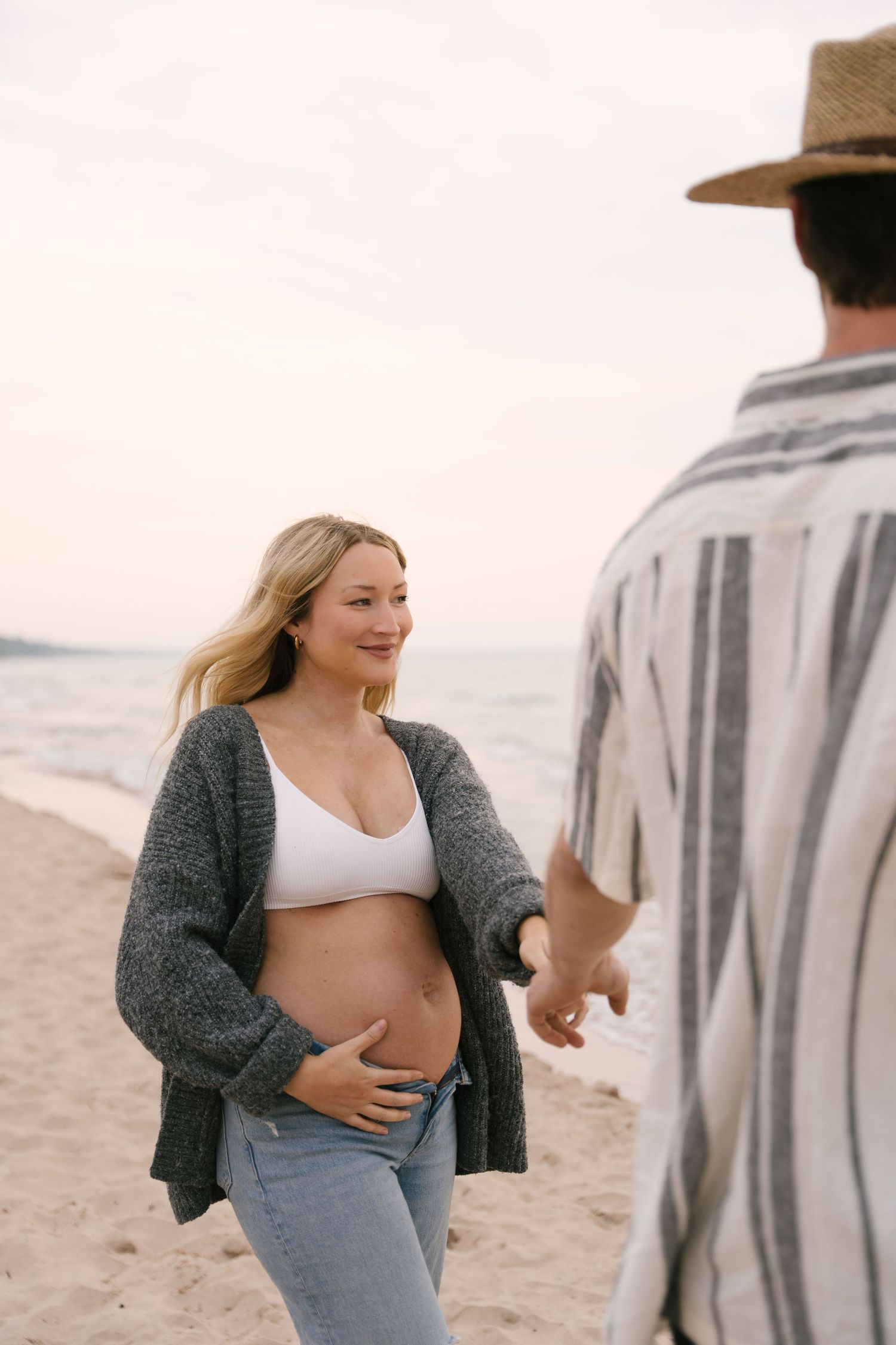 Smiling pregnant woman in gray cardigan and white bralette reaching for partners hand on beach.