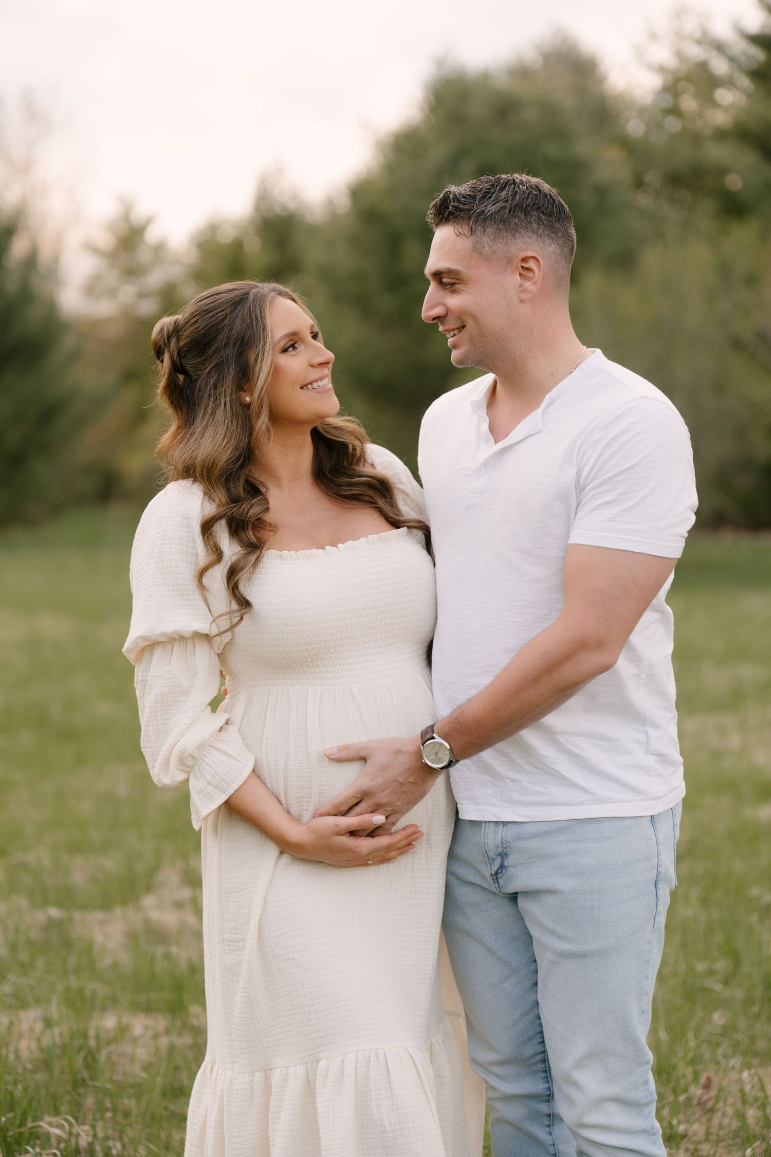 Smiling couple in white outfits cradling pregnant belly in a sunny green field.