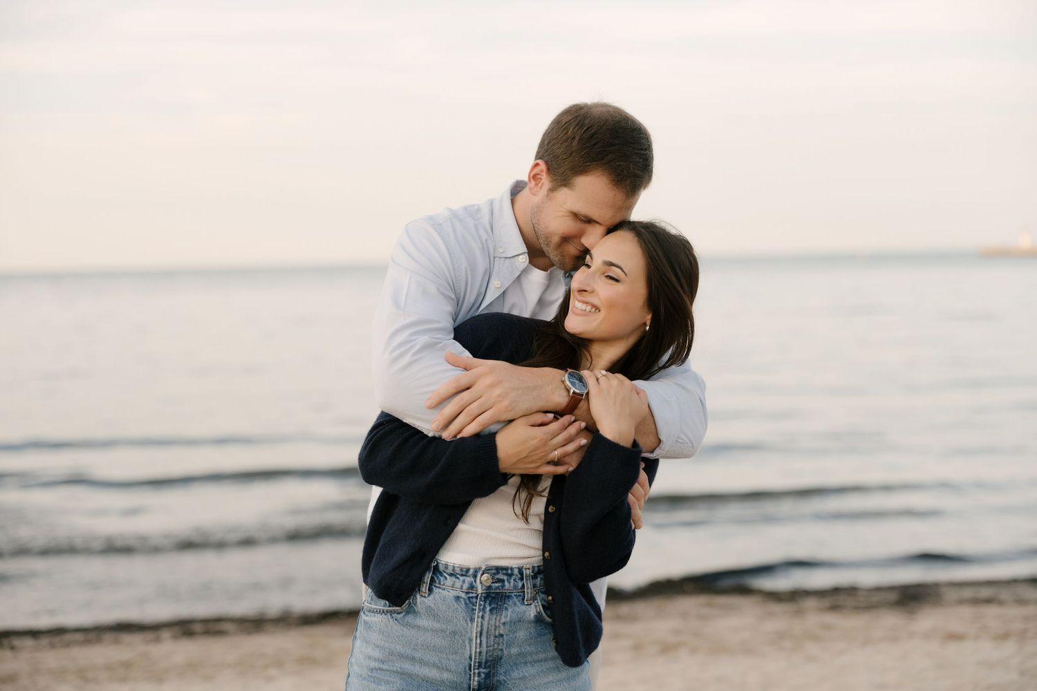 Man embracing woman from behind on beach at sunset, both smiling with ocean waves in background.