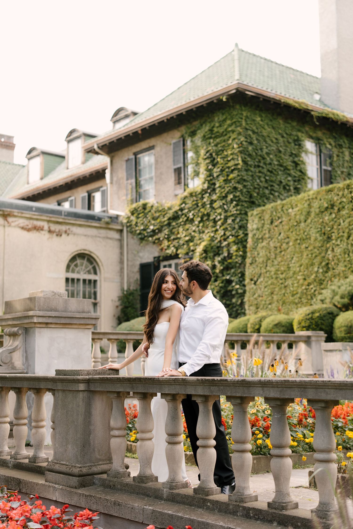 Romantic couple standing on stone balustrade with ivy-covered mansion and colorful garden.