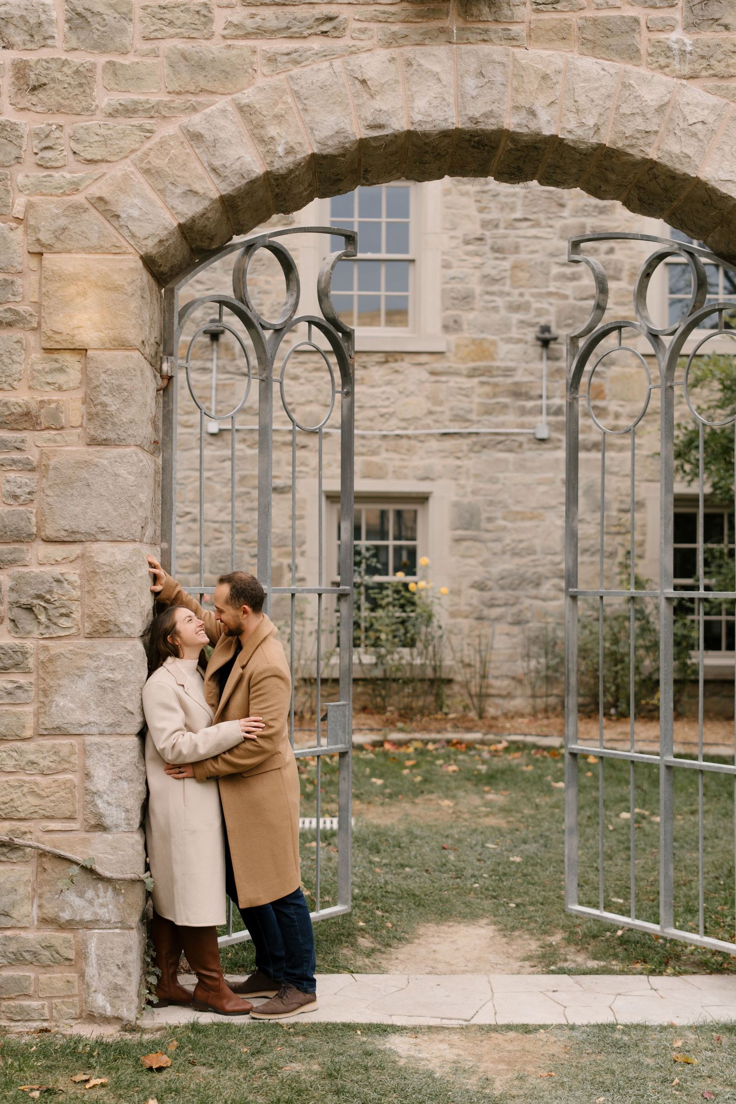 Couple sharing a romantic moment under a stone archway with ornate iron gates in autumn garden.