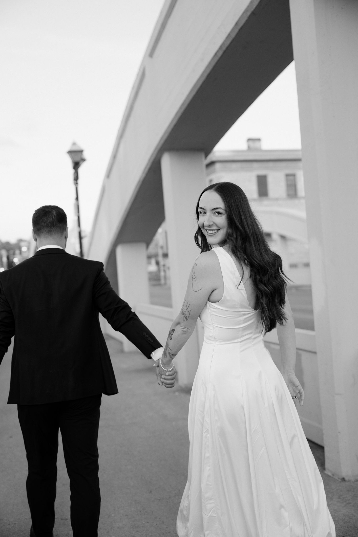 Black and white photo of bride looking back smiling while holding grooms hand near bridge.