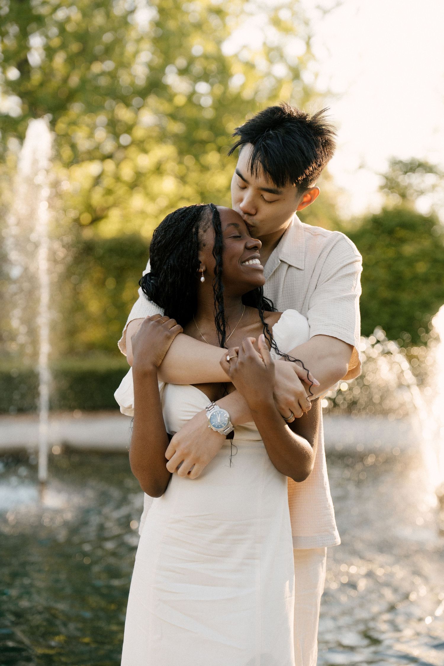 Man kissing woman on forehead while embracing her from behind near a garden fountain at sunset.