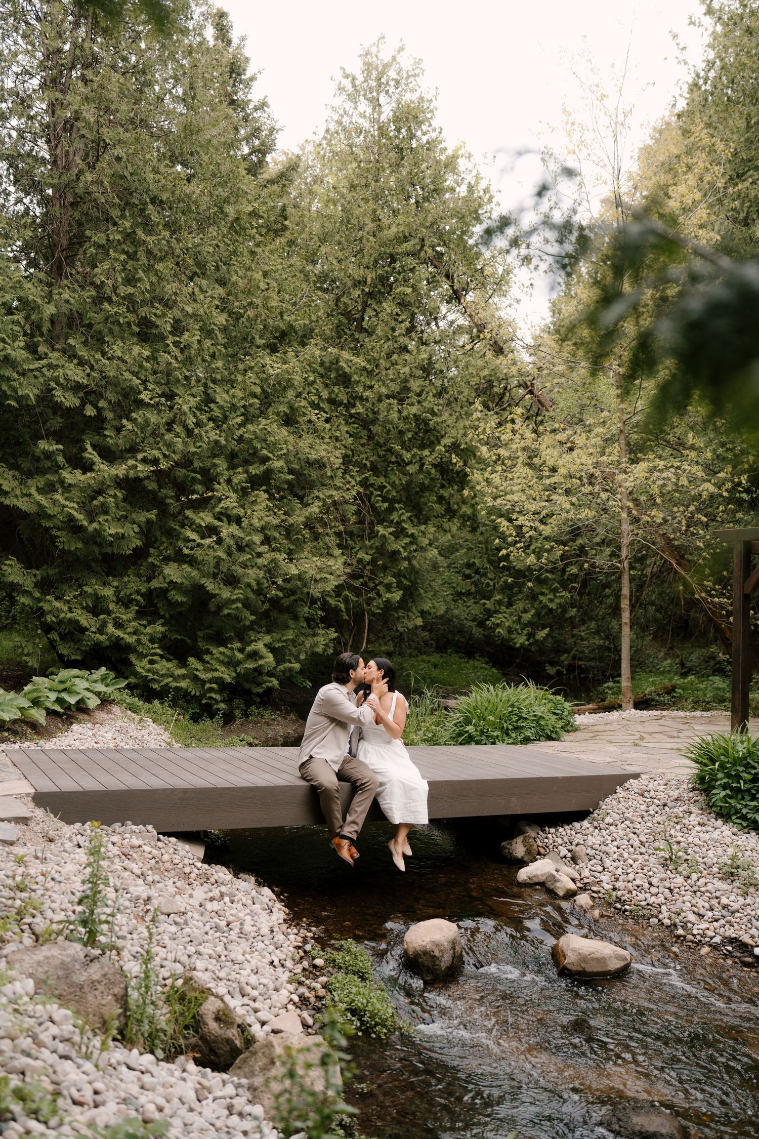 Couple sitting and kissing on a wooden bridge over a flowing stream in a serene green forest setting.