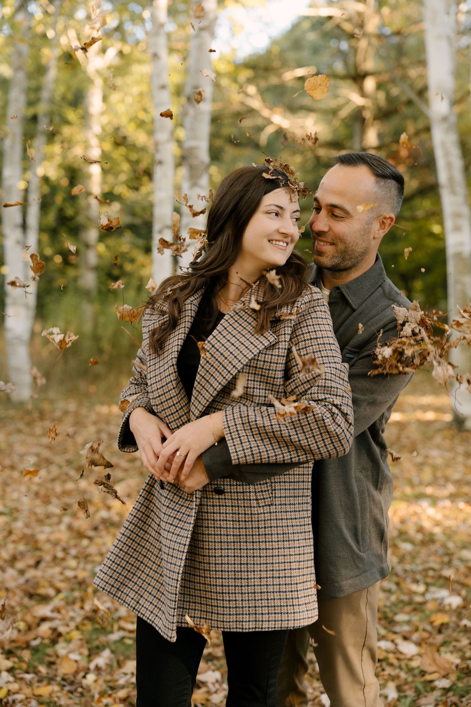 Couple embracing as fall leaves cascade around them in a sunlit birch forest.