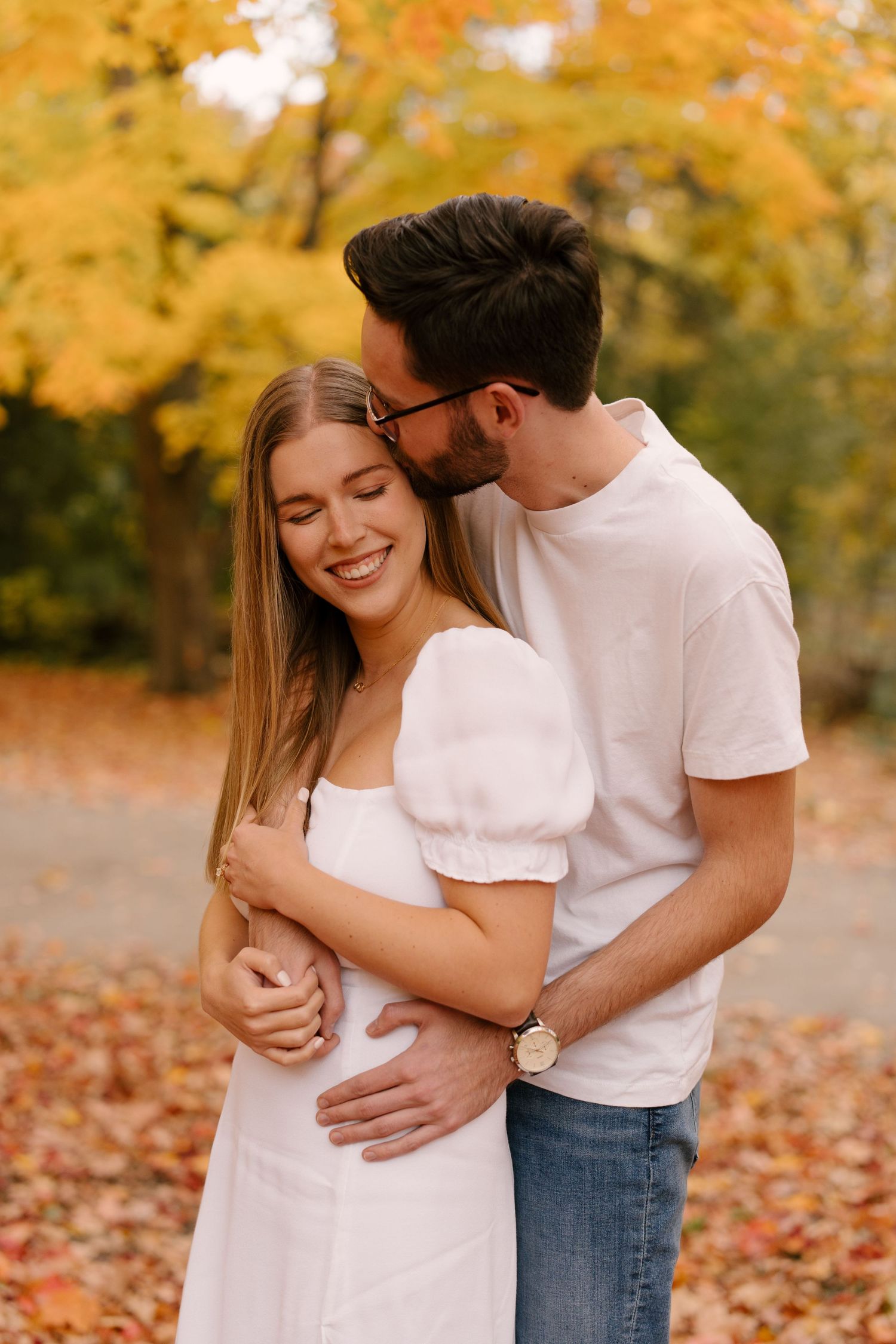 A happy couple embraces in an autumn park, the man kissing the woman's cheek as she smiles.