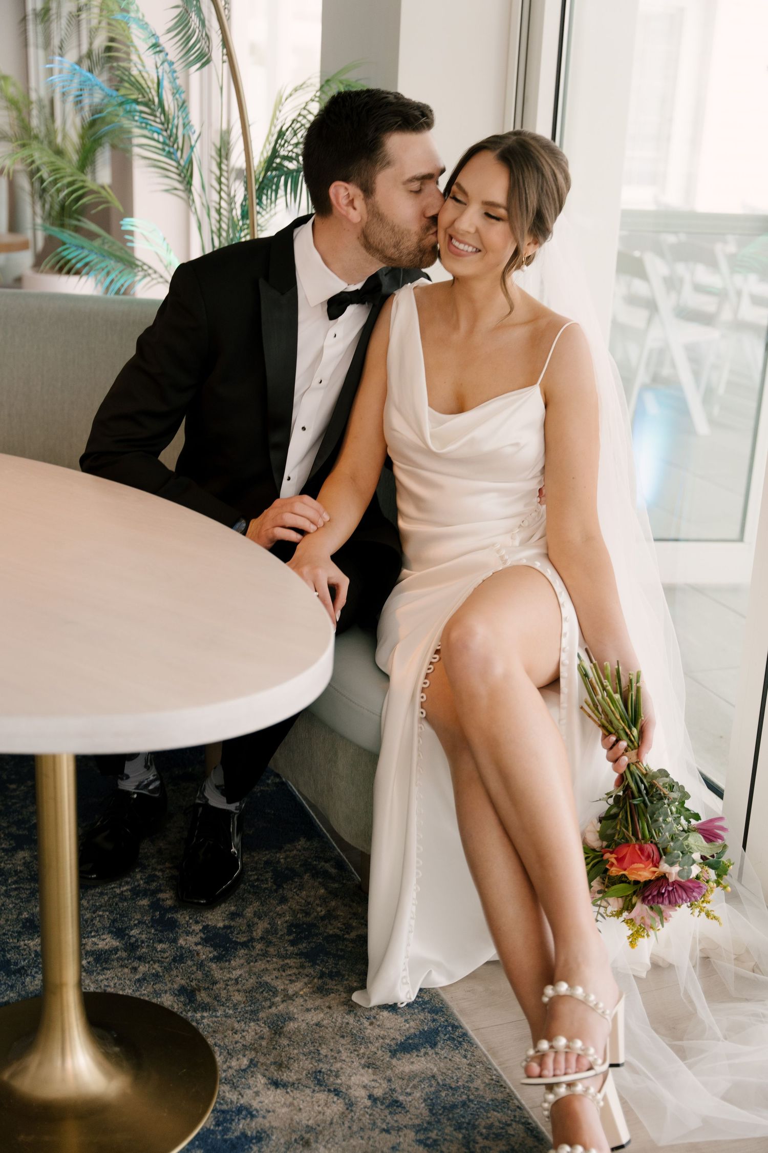 Groom kisses bride on cheek as they sit together, bride holding vibrant floral bouquet indoors.