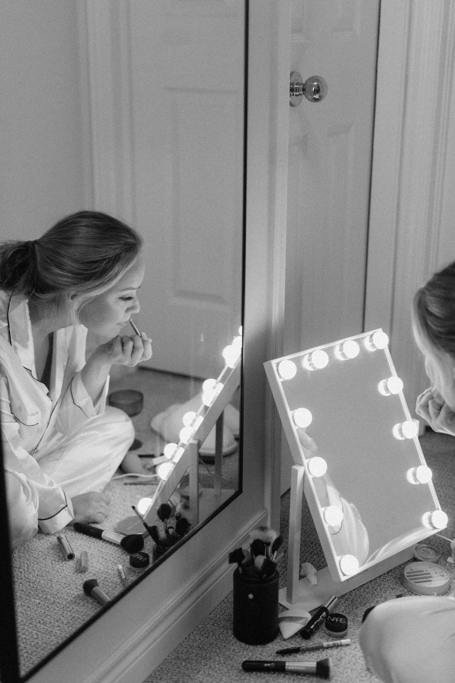 Bride sitting on floor applying makeup using a lighted vanity mirror in black and white photo.