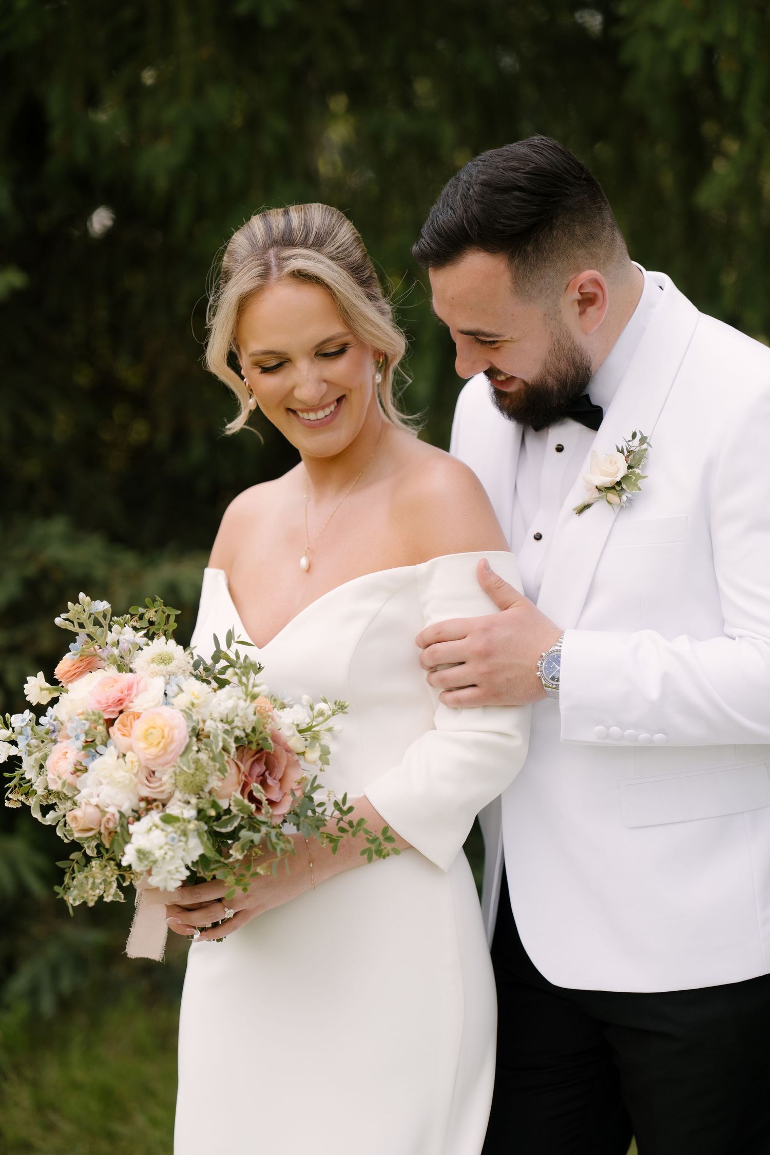 Smiling bride in off-shoulder white gown and groom in white tuxedo sharing a joyful moment with pastel bouquet.