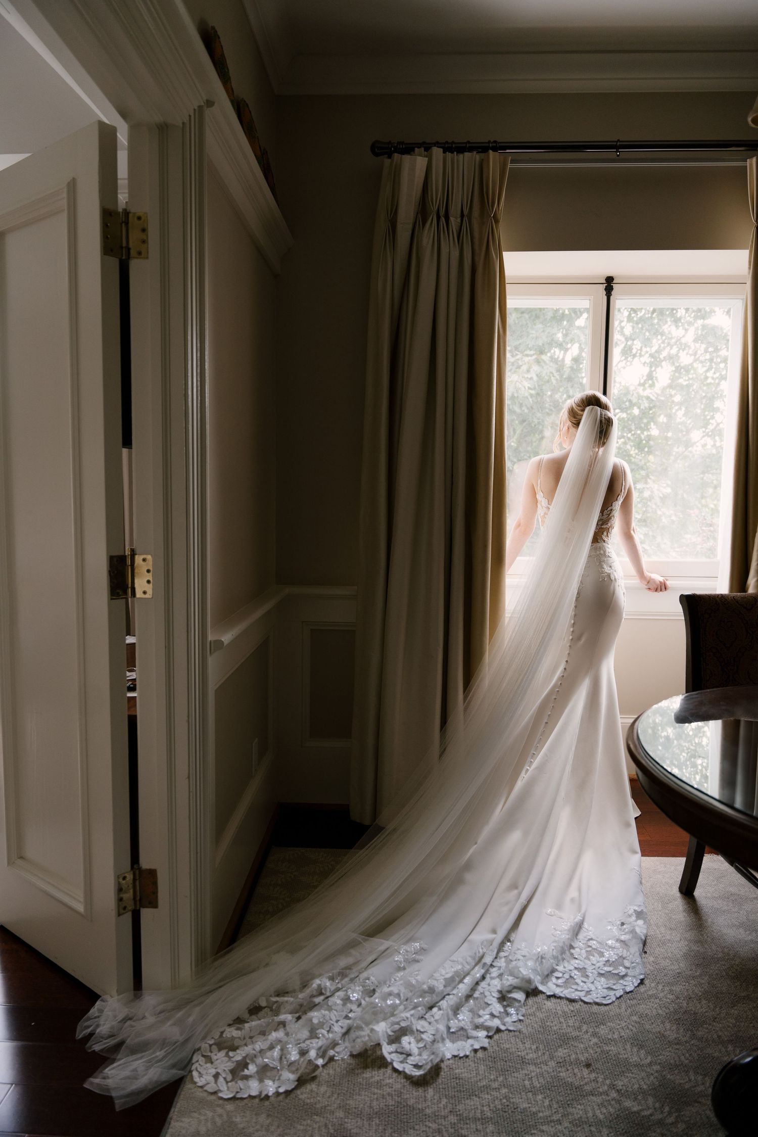 Bride in a fitted lace wedding gown with a long cathedral veil stands gazing out a window in an elegant Langdan Hall room.