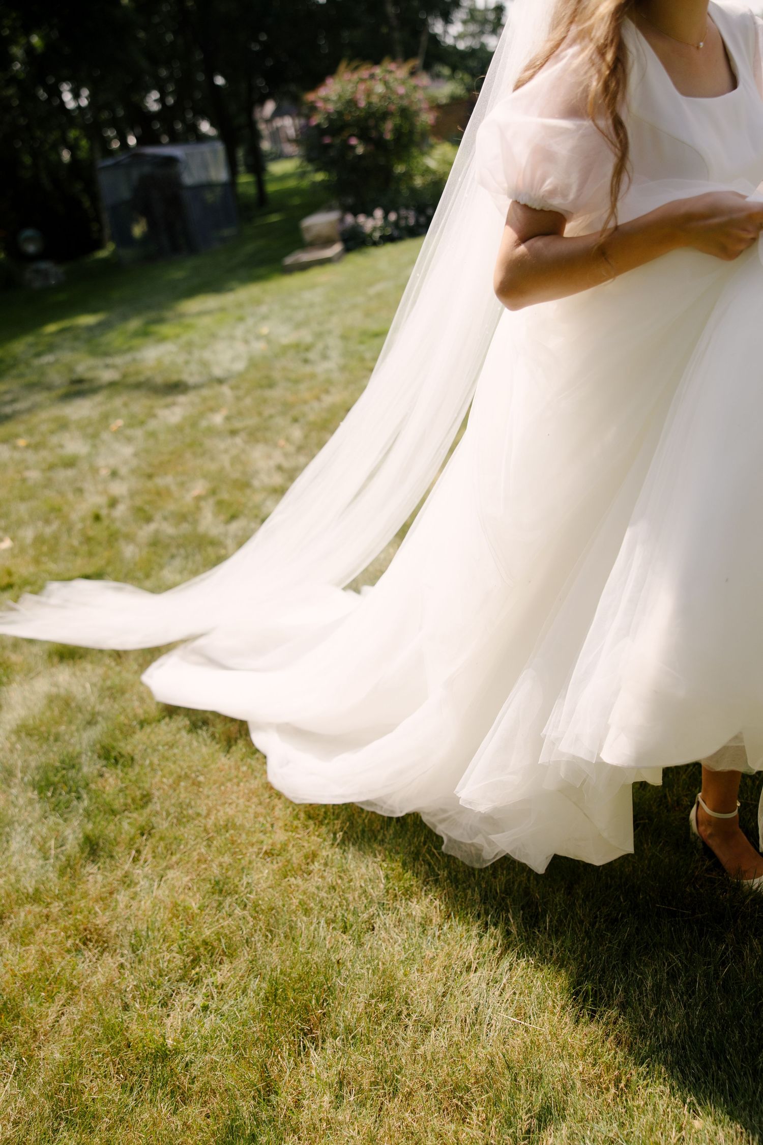 Bride walking on grass in flowing white tulle wedding gown with long veil train in sunlit garden.
