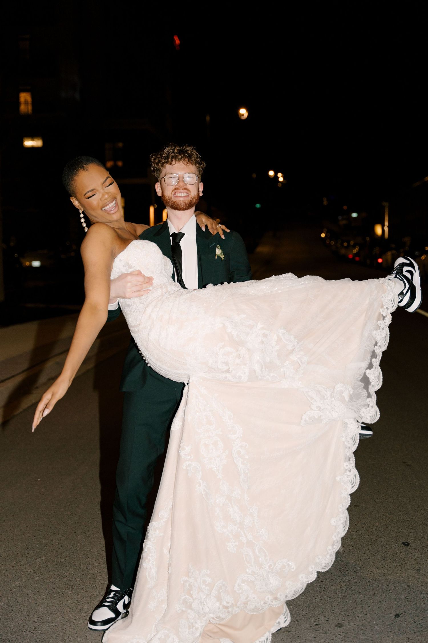Groom in dark suit carrying laughing bride in white lace wedding gown outside at night at Tapestry Hall.