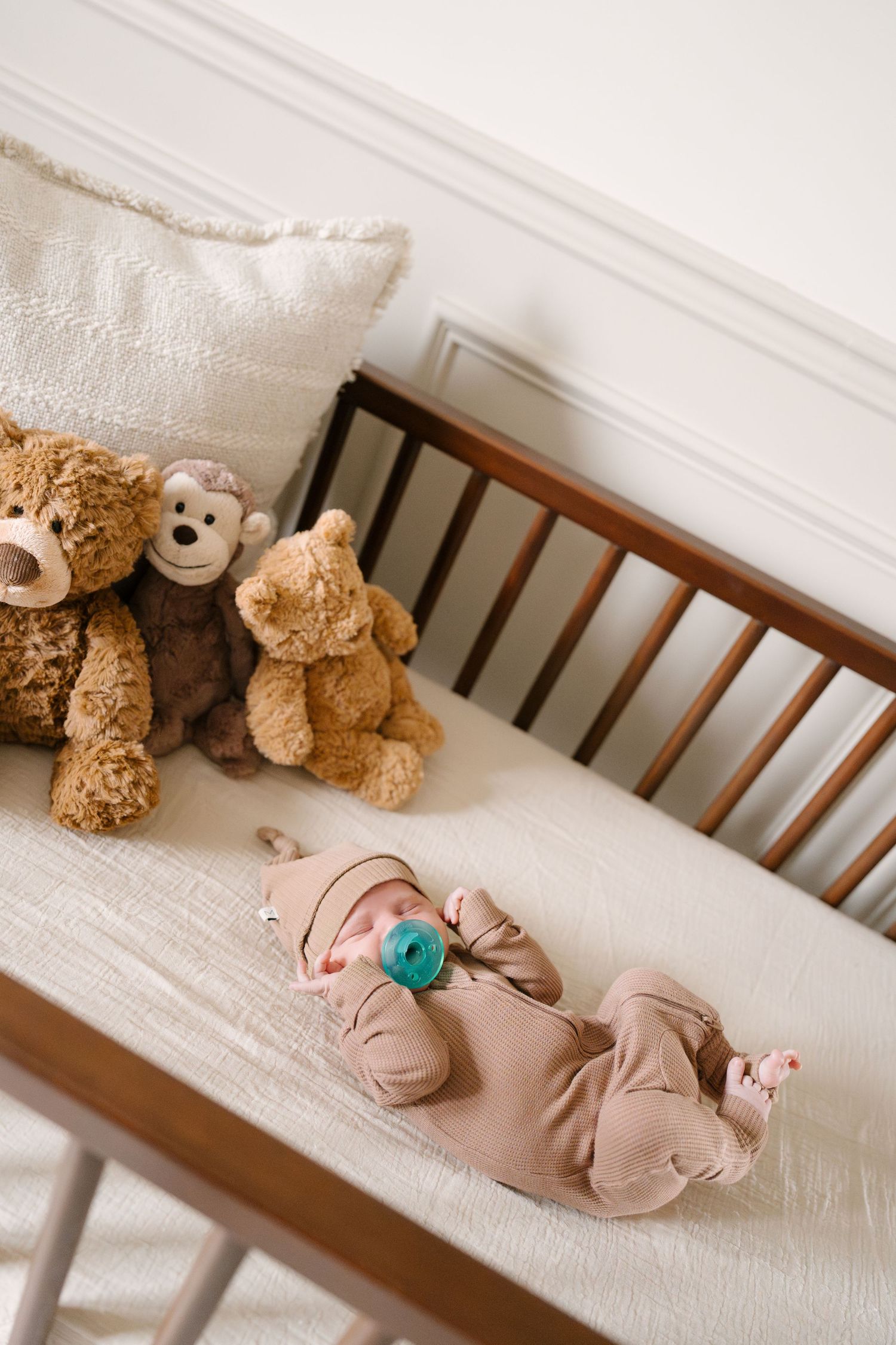 Newborn baby with pacifier sleeps in a wooden crib beside three teddy bears on white bedding.