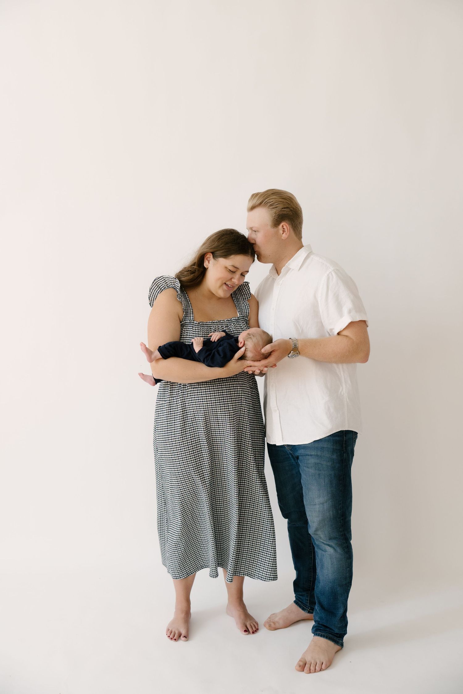 Happy parents in a bright studio lovingly gaze at their newborn baby dressed in navy.