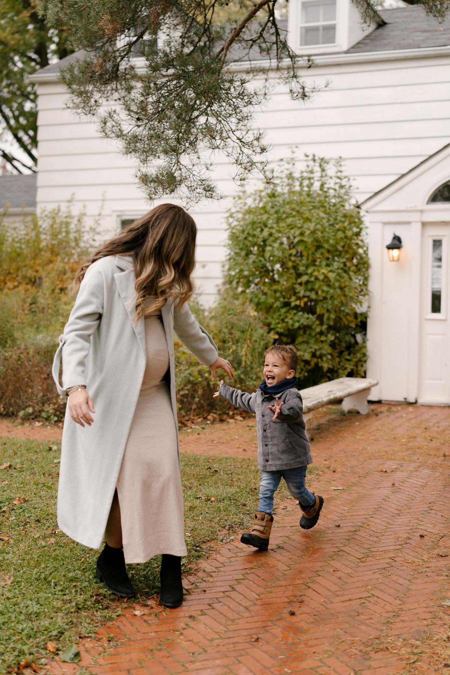 Pregnant mother in grey coat holding hands with running toddler on brick path outdoors.