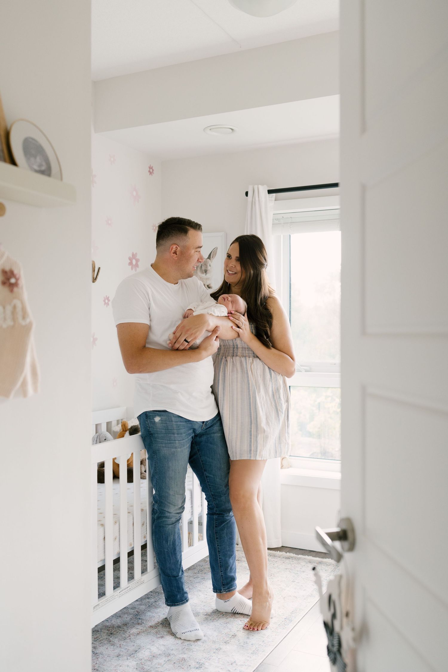 New parents smiling at newborn baby in bright white nursery with pink floral wallpaper.