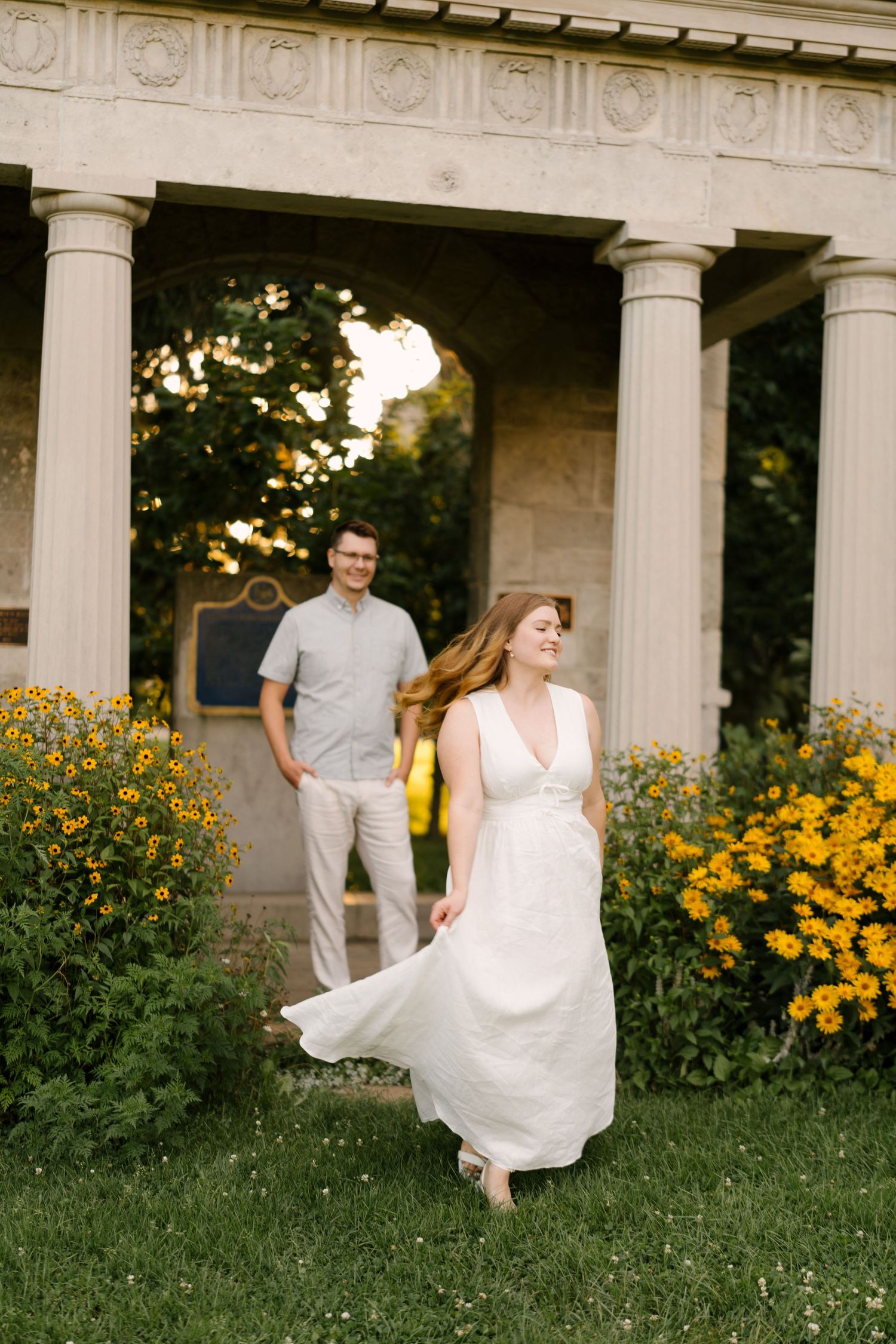 Woman in white dress twirling near stone columns with yellow flowers, man smiling in background.