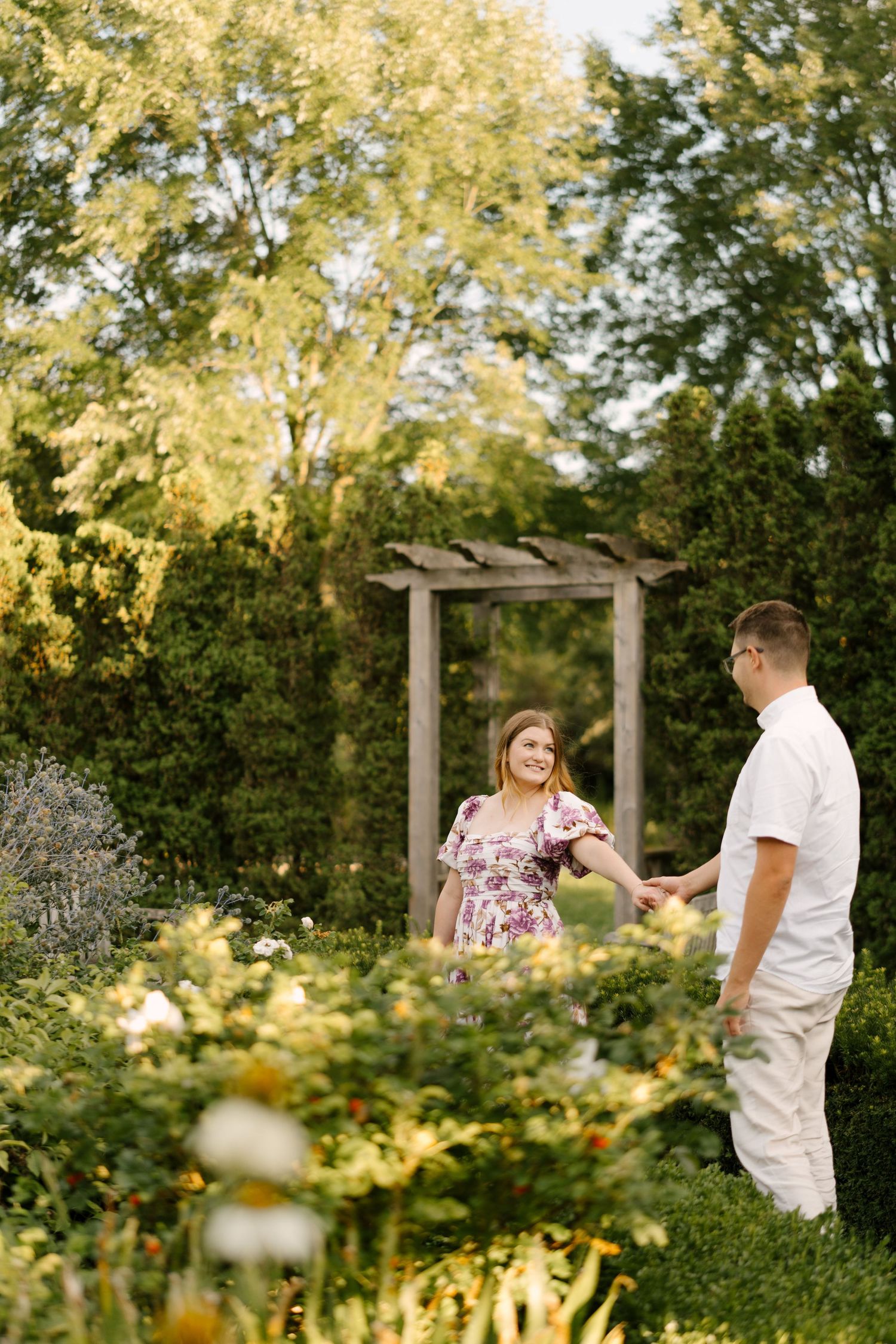 Smiling couple holding hands in blooming garden with wooden pergola arch at dusk.