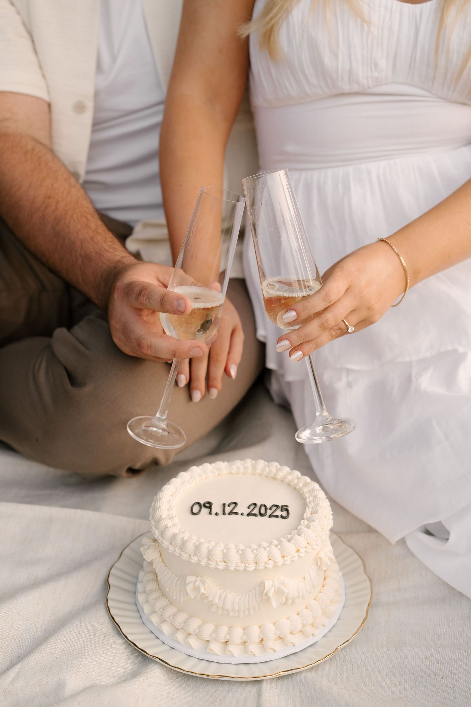 Couple holding champagne flutes beside a white two-tier cake inscribed with 09.12.2025 outdoors.