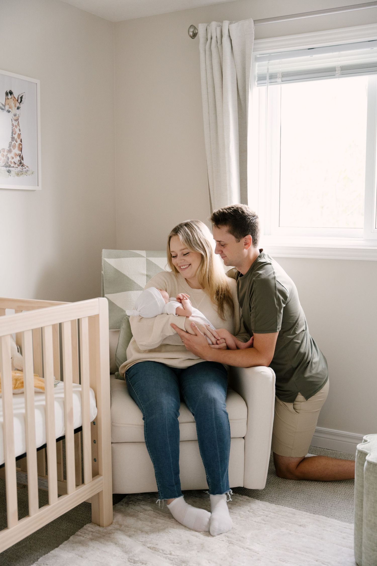 New parents sitting together in nursery chair by window lovingly gazing at their newborn baby.