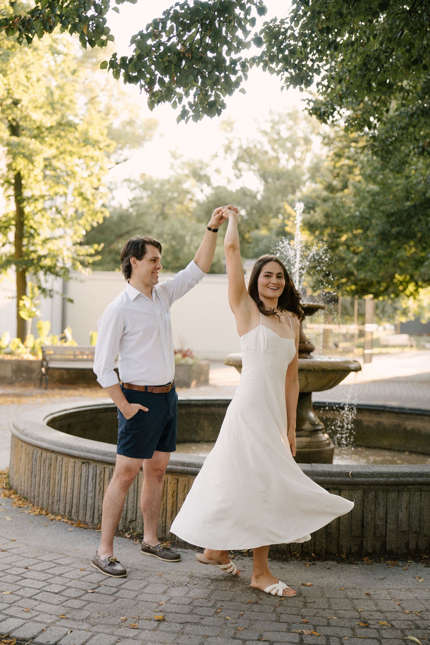 Couple dancing by a fountain in a sunny park, woman in white dress twirling.