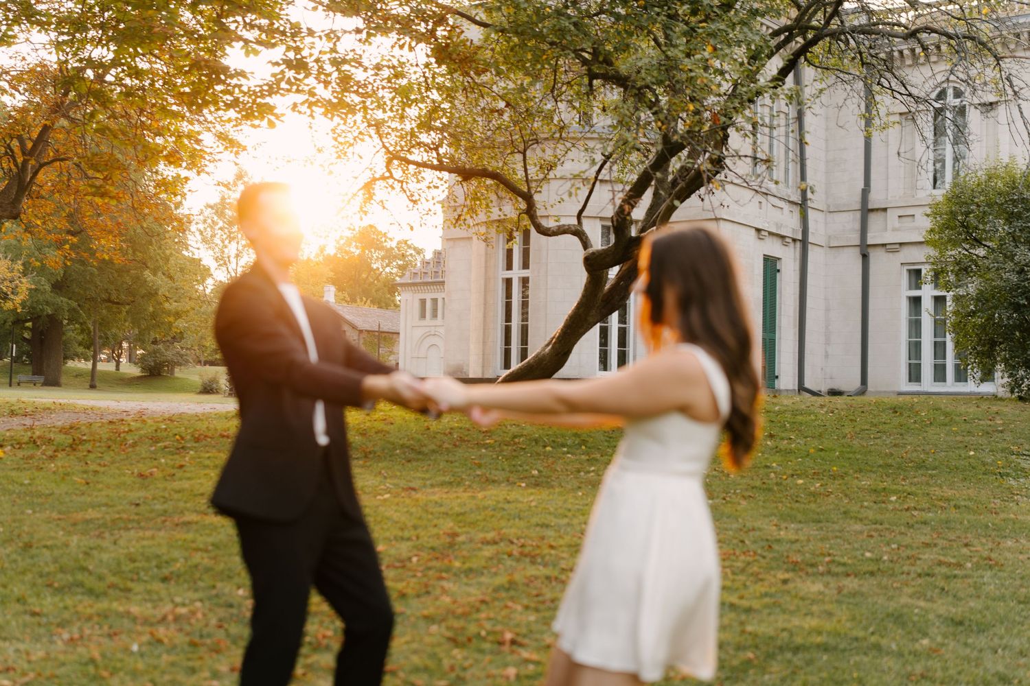 Couple holding hands spinning joyfully on lawn at golden sunset with large estate building behind them.